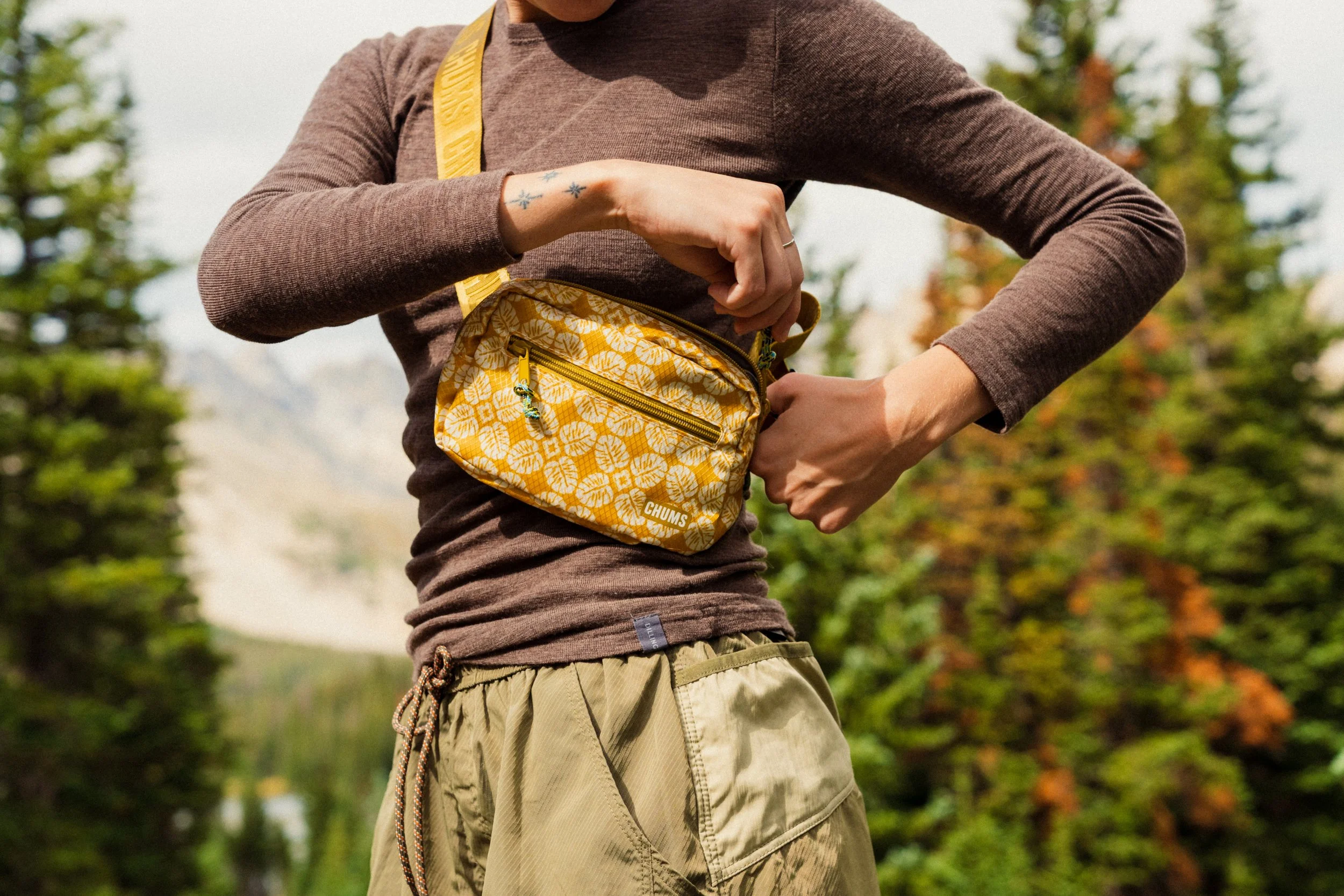 Person wearing a brown long-sleeve shirt and beige cargo shorts, holding a yellow patterned small bag with a zipper in an outdoor forest setting.