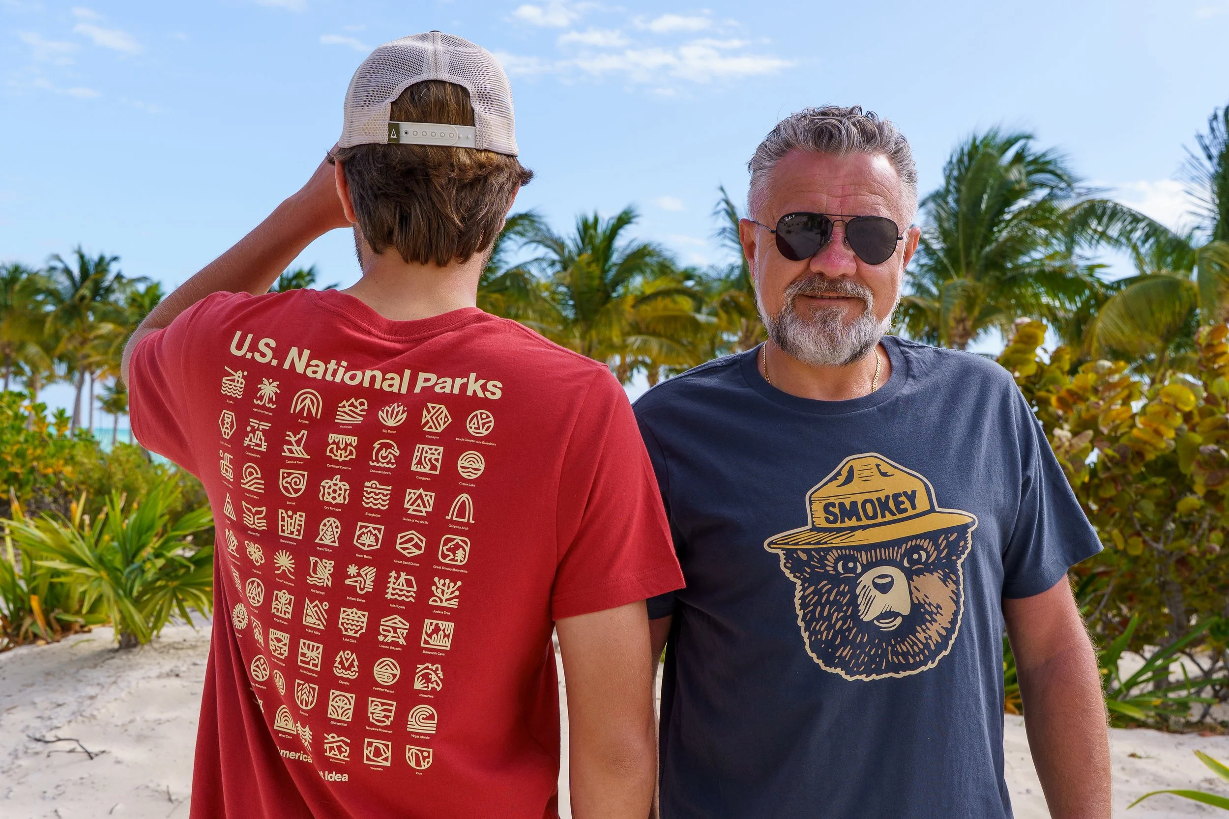 Two men standing outdoors on a sunny day with palm trees in the background. One man is wearing a red U.S. National Parks t-shirt, and the other man is wearing sunglasses and a blue graphic t-shirt with a bear image and the word 'Smokey.' The man in the red shirt is facing away from the camera, and the man in the blue shirt is facing forward.