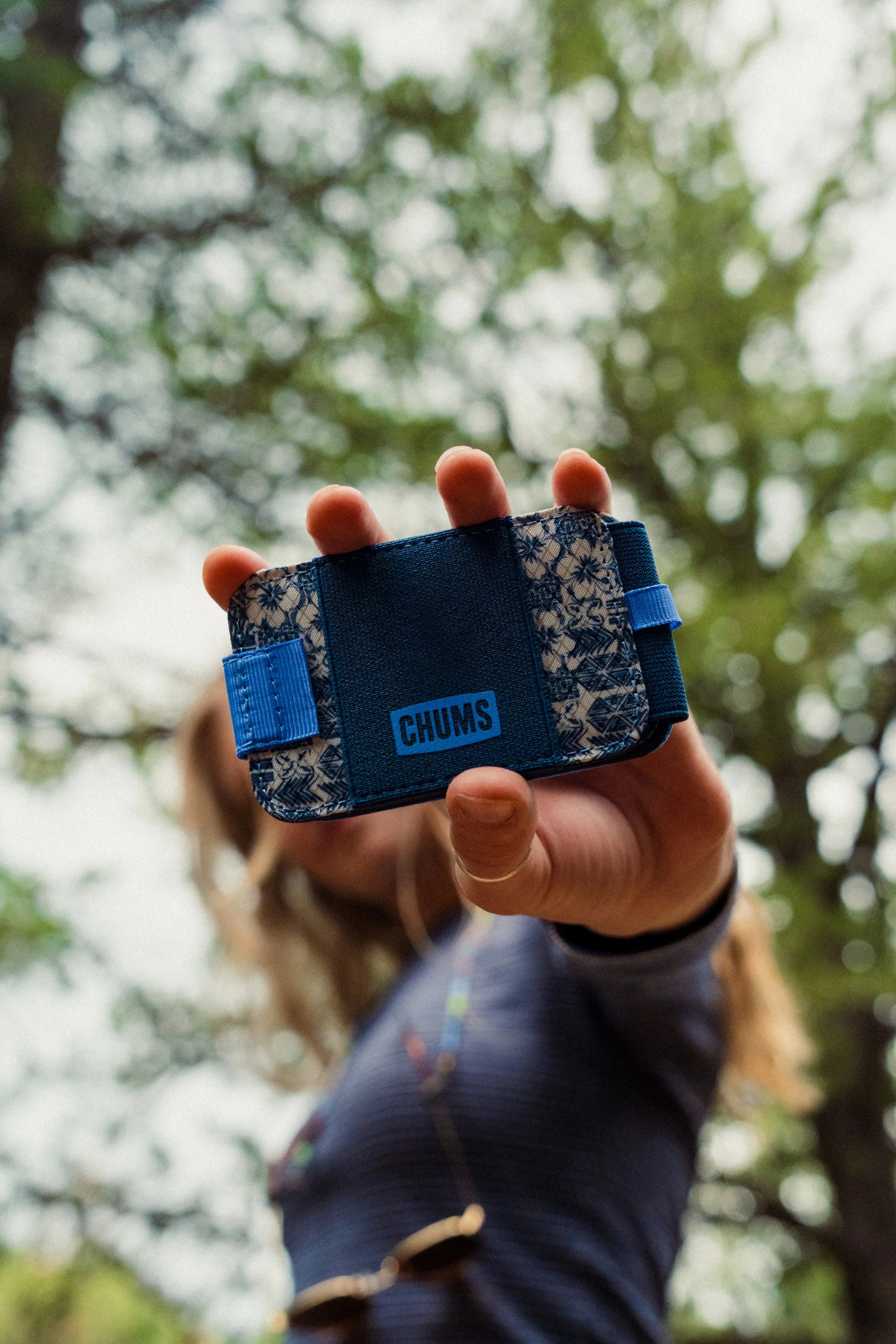Person holding a small, patterned blue wallet with the brand name CHUMS, outdoors with trees in the background.