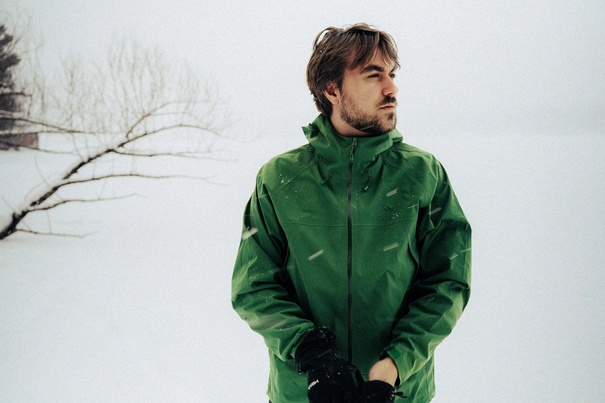 A young man with brown hair and a beard wearing a green jacket standing outdoors in snowfall during daytime.