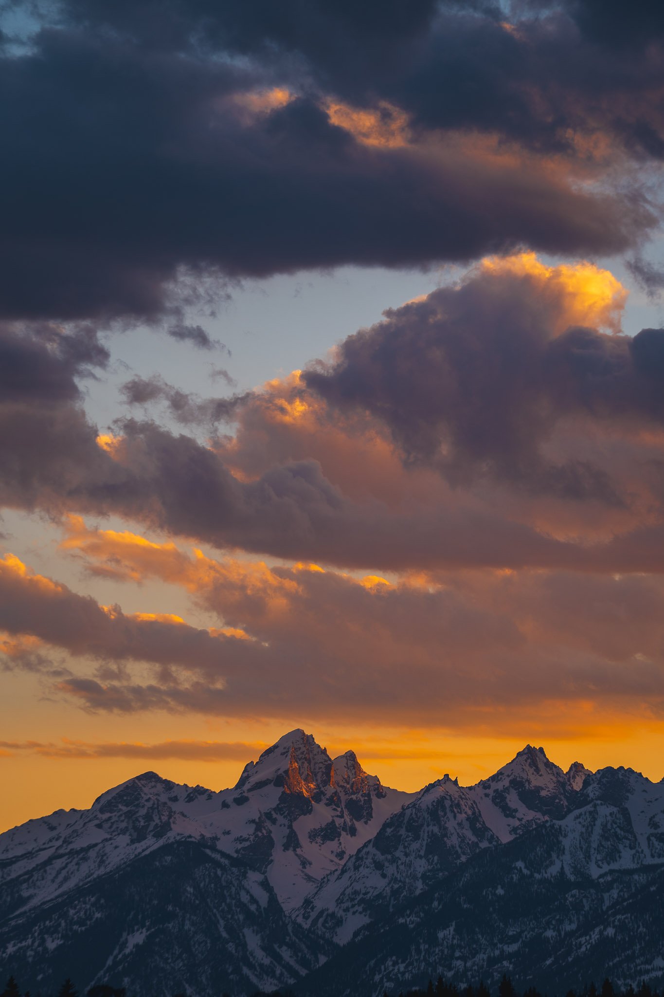 Snow-capped mountains under a colorful sunset sky with dark and illuminated clouds.