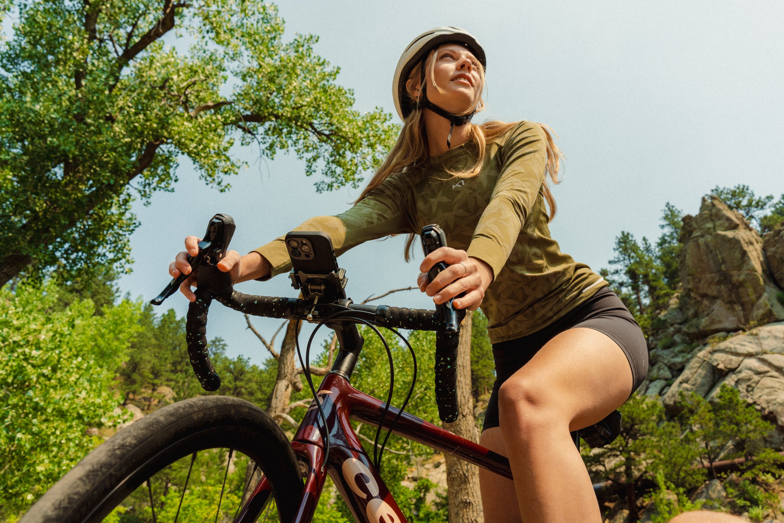 Woman in outdoor gear riding a mountain bike through a forested area with rocks and trees, wearing a helmet and athletic clothing, looking into the distance.