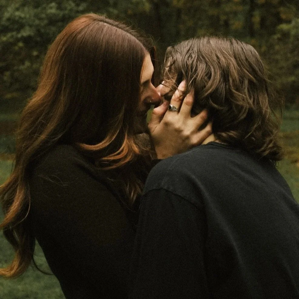 A woman with long wavy brown hair holding a man with shoulder-length curly hair face-to-face in an outdoor setting, appear to be about to kiss.