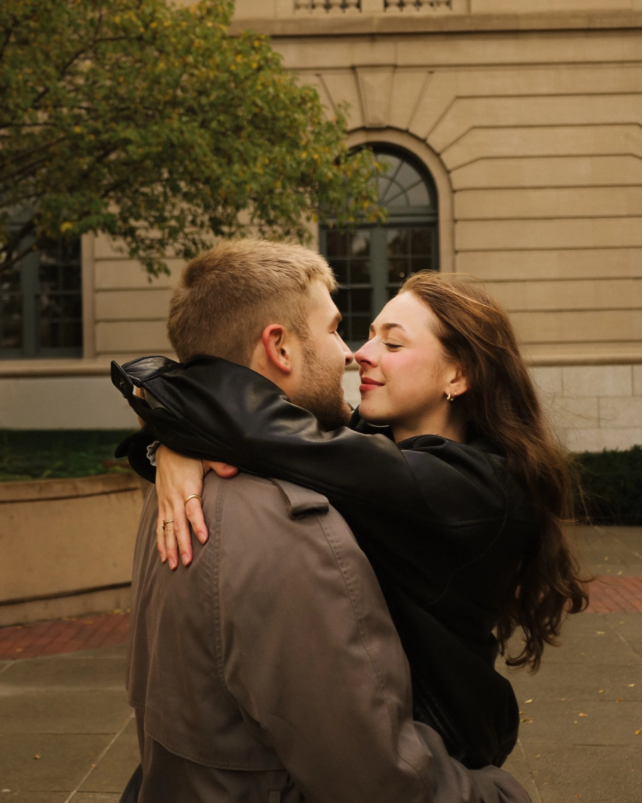 A couple embracing each other outdoors with their faces close together, eyes closed, in front of a beige building with arched windows and trees.