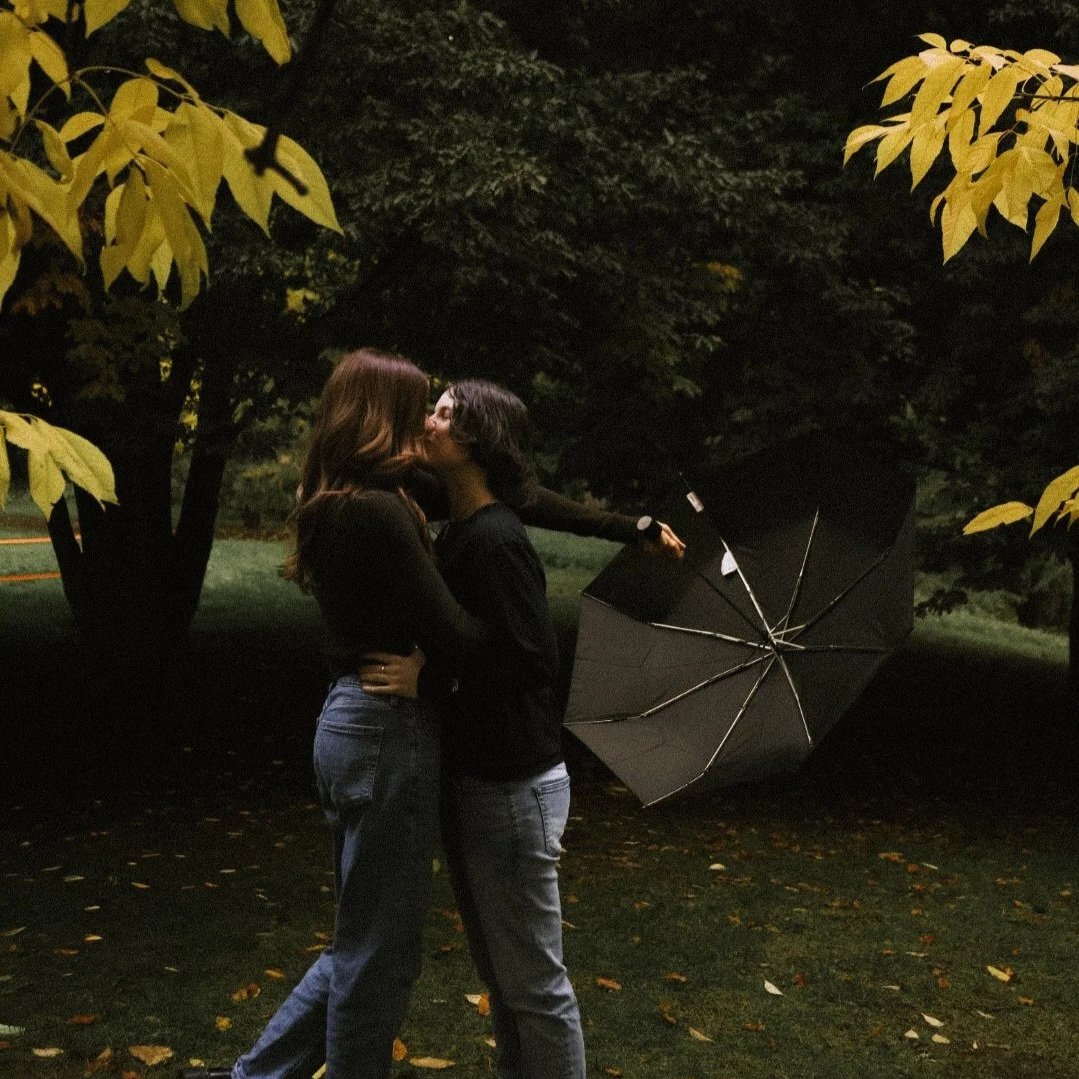 Two women kissing outdoors at night, standing under trees with yellow leaves, with a black umbrella nearby.