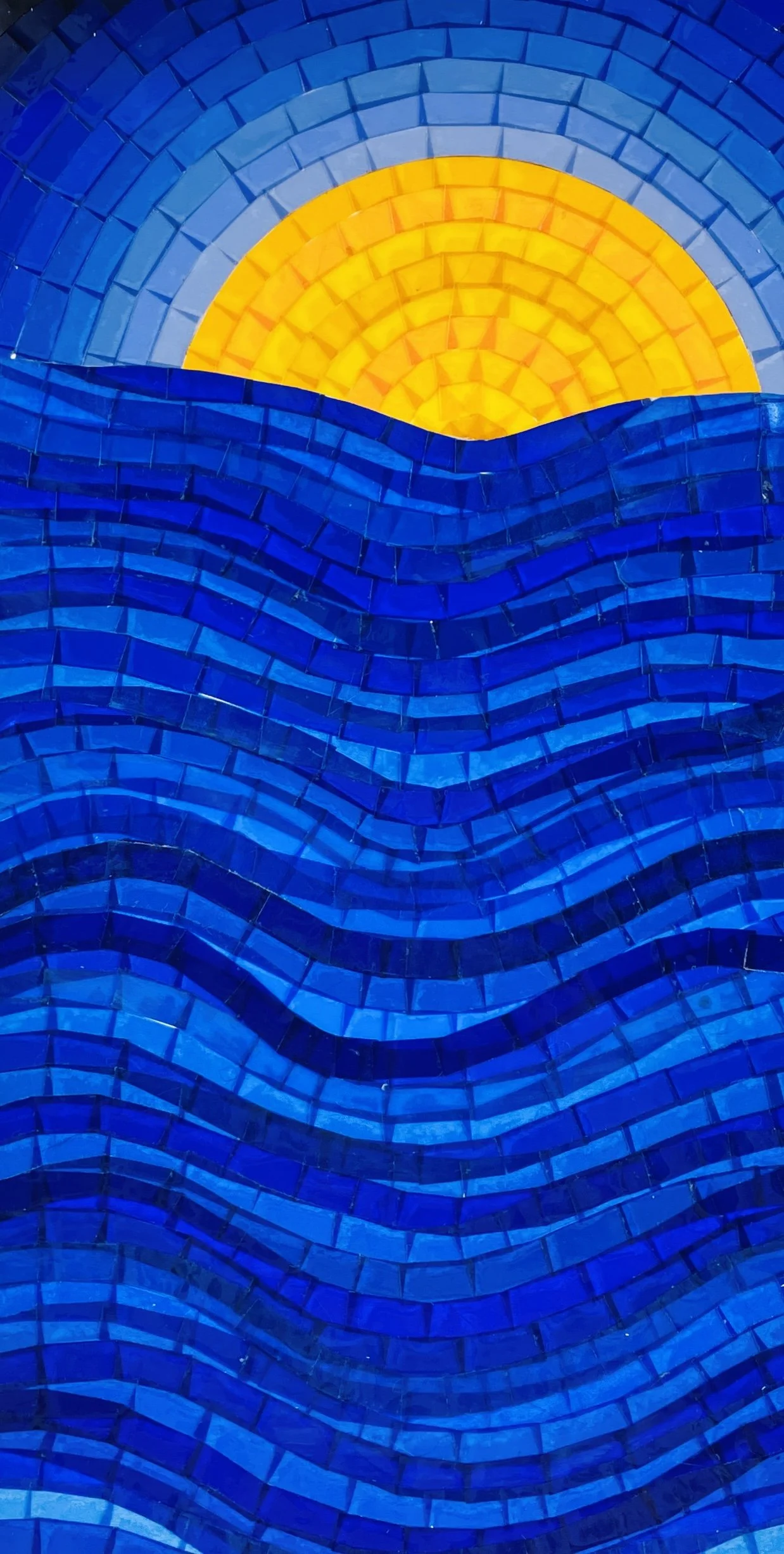 *"Sun Over Sea" (2026). 5.5x10.5". A narrow 5.5x10.5" mosaic made out of small segments of translucent plastic. A yellow sun half-rises on the horizon above a swirling ocean in rich shades of blue. The medium makes the piece look almost liquid.