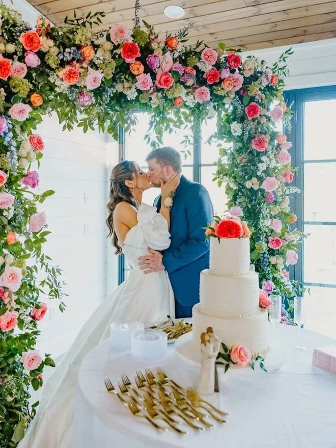 A bride and groom kiss under a large floral arch with pink, red, purple, and white flowers at their wedding reception.