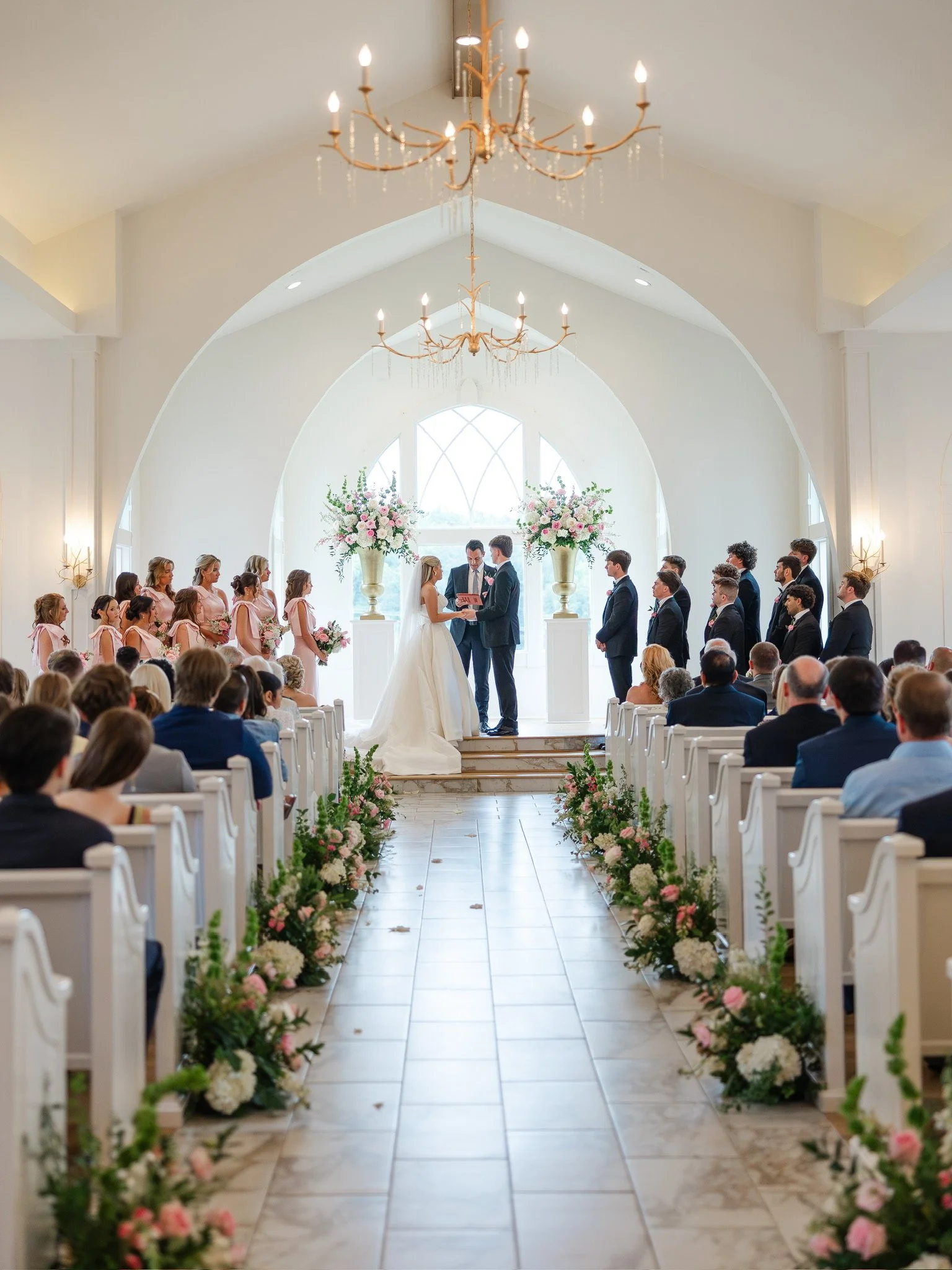 A wedding ceremony inside a bright, white chapel with high, vaulted ceilings and large windows. The bride and groom stand at the altar, exchanging vows, with bridesmaids in pink dresses on the left and groomsmen in dark suits on the right. Floral arr