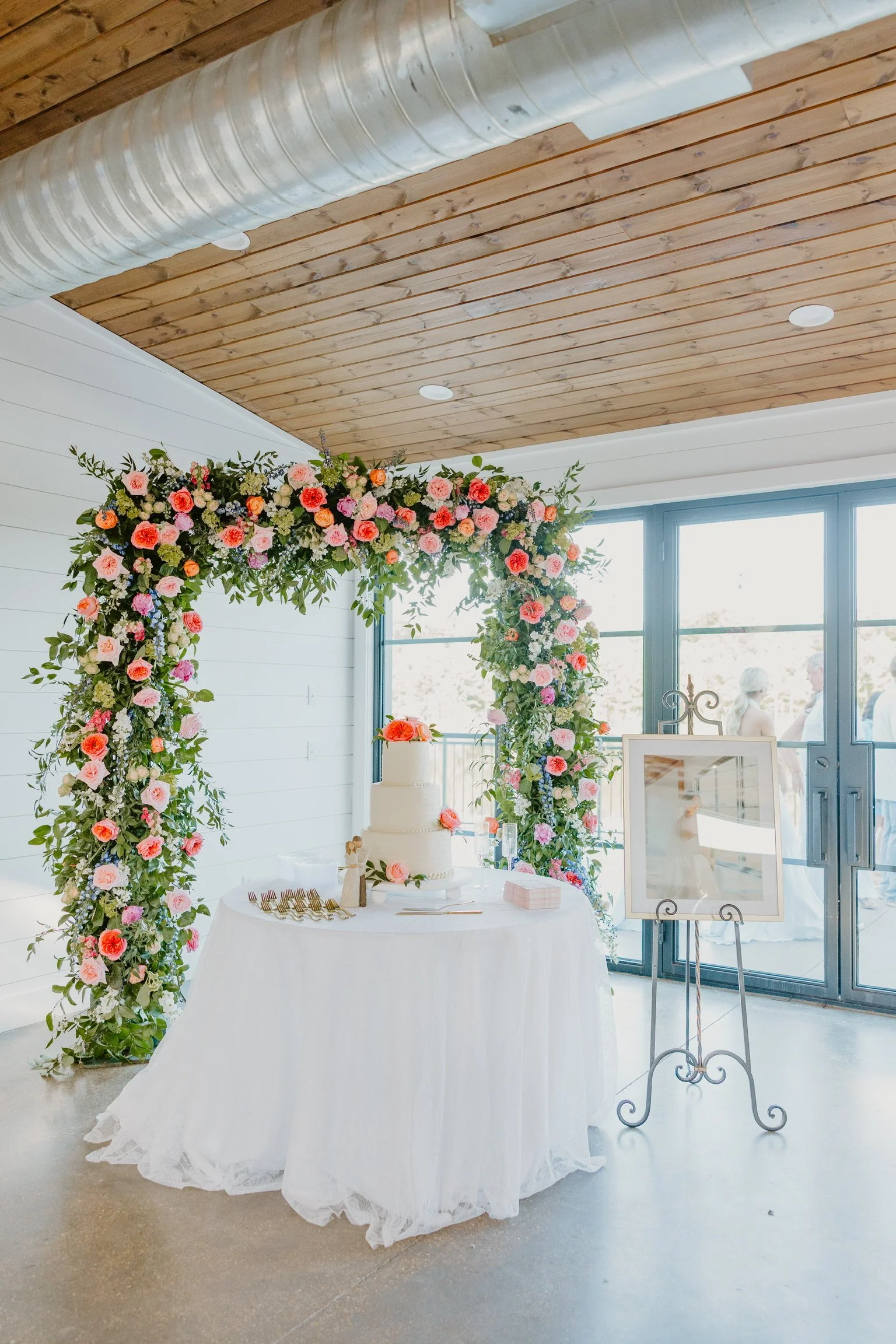 A decorated wedding or event reception area with a white tablecloth, a tiered cake, and a floral arch made of pink, orange, white, and purple flowers with green foliage.
