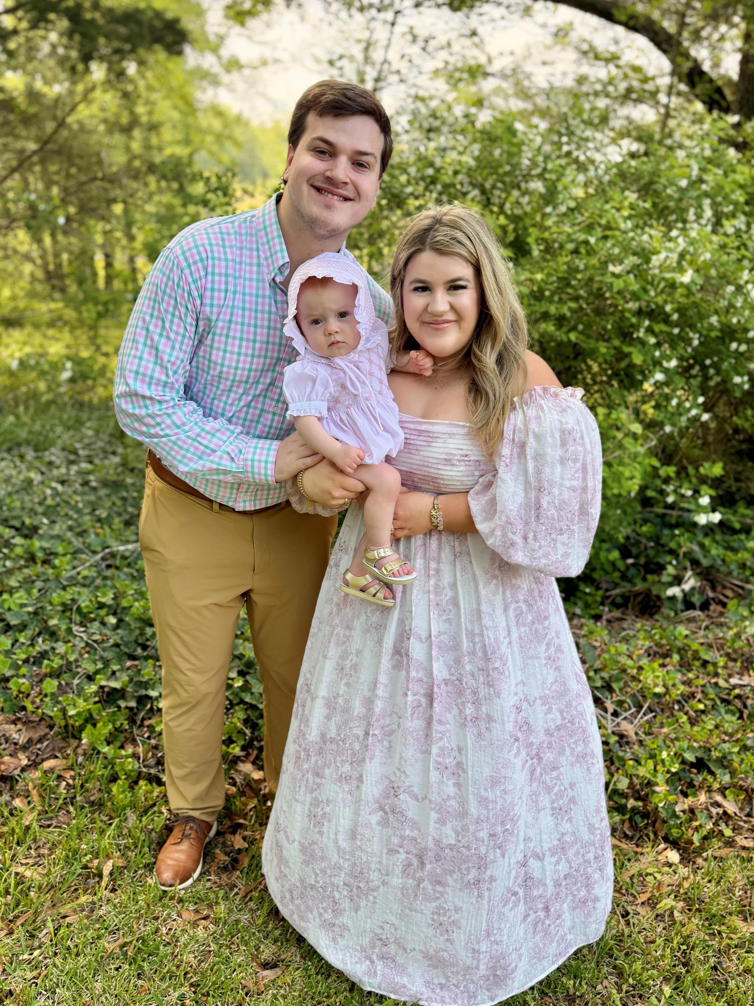 Family photo of a man, woman, and baby girl outdoors surrounded by green trees and grass during daytime.