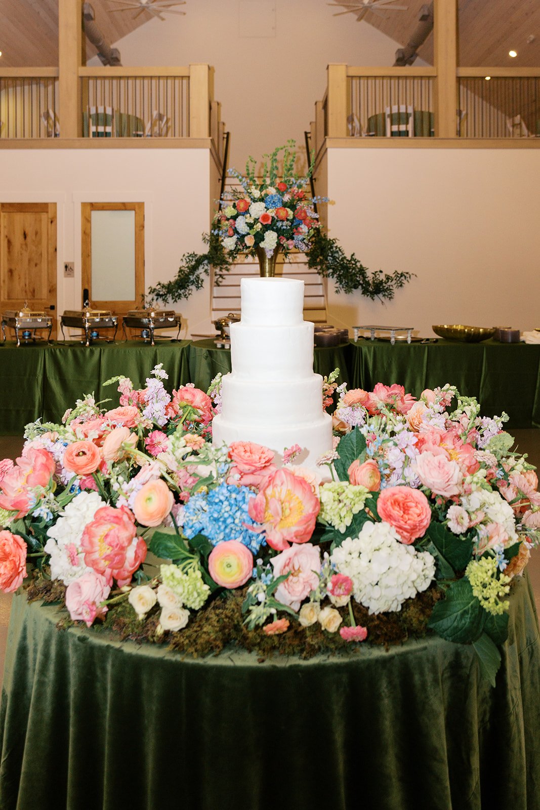 A white, three-tiered wedding cake on a table with a green tablecloth, surrounded by pink and white flowers. There is a large floral arrangement on top of the cake and another large flower arrangement in the background, with a buffet table behind. Th