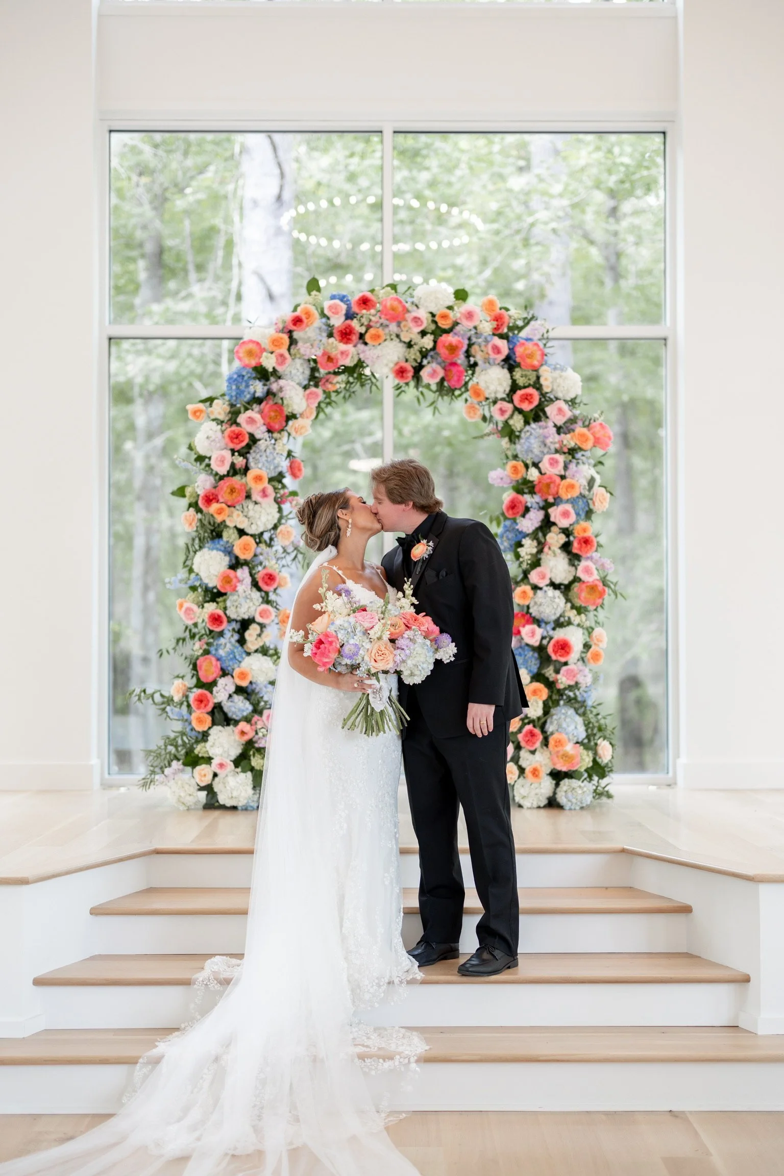 A bride and groom kissing in front of a large floral wedding arch with pink, white, purple, and blue flowers, inside a modern building with large windows showing green trees outside.