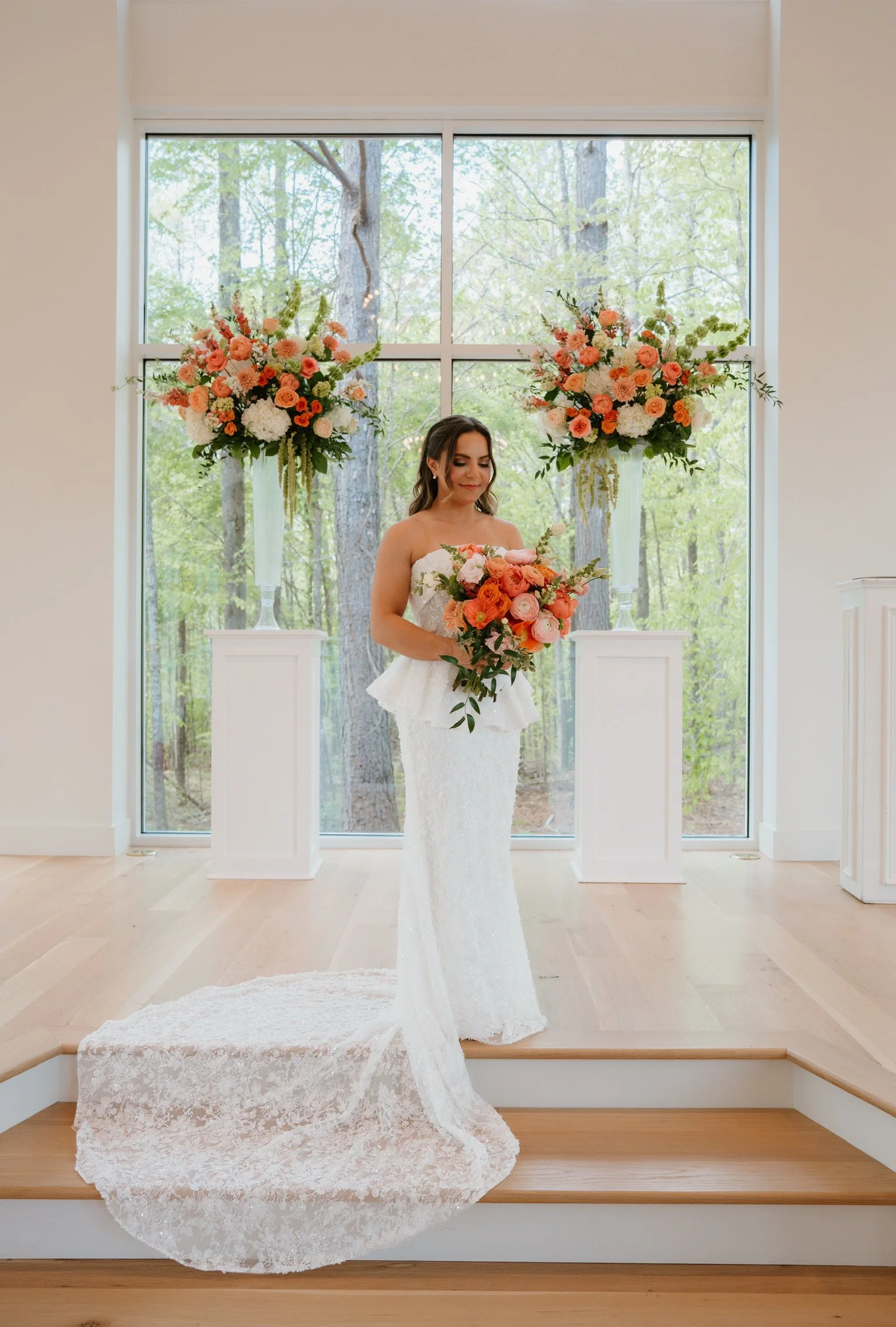 A bride in a white lace wedding dress holding a bouquet of flowers standing on a wooden staircase in front of a large window with trees outside, with two tall floral arrangements on white pedestals flanking her.