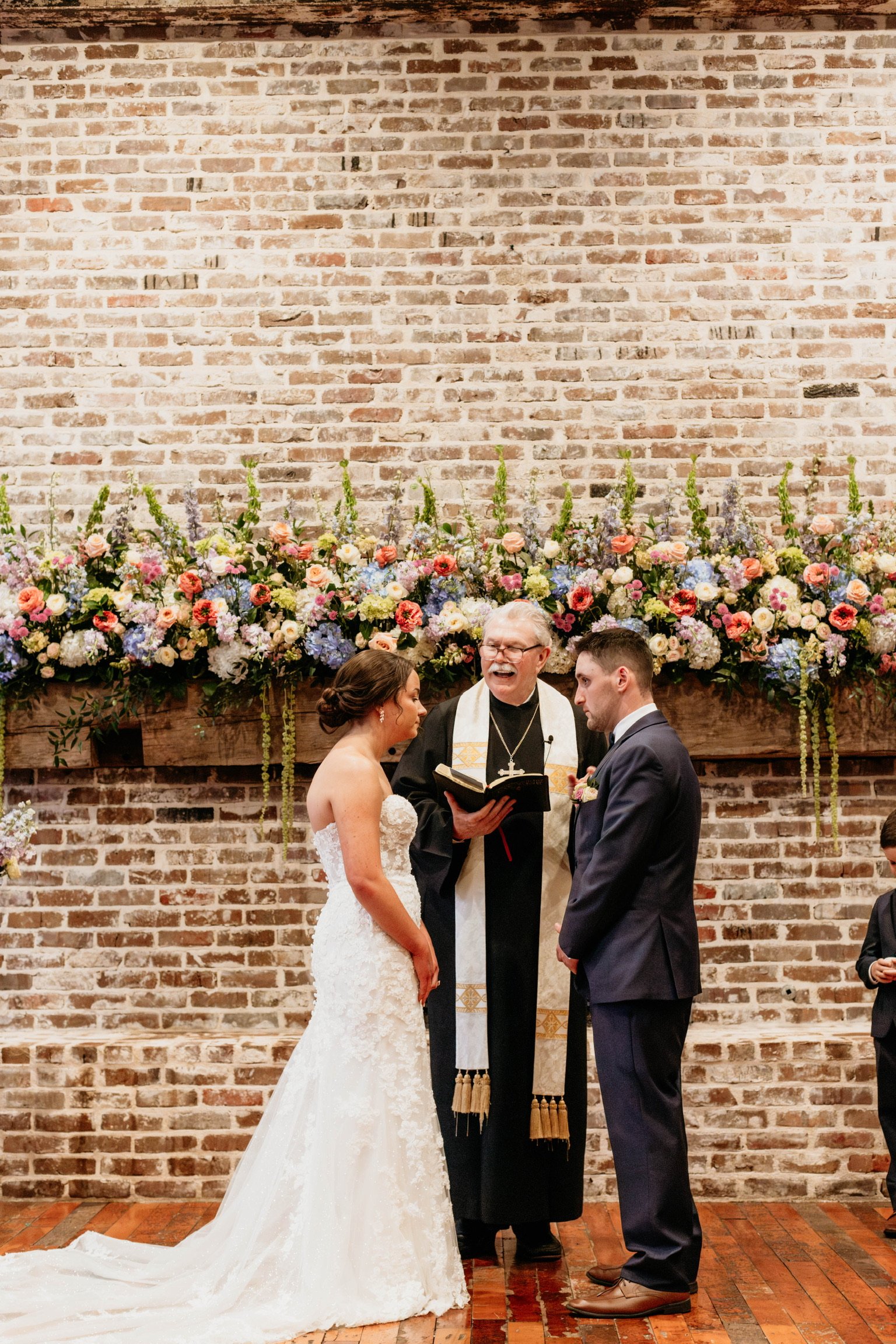 A bride and groom standing face to face in front of a brick wall with a large floral arrangement, officiant holding a book, during a wedding ceremony.
