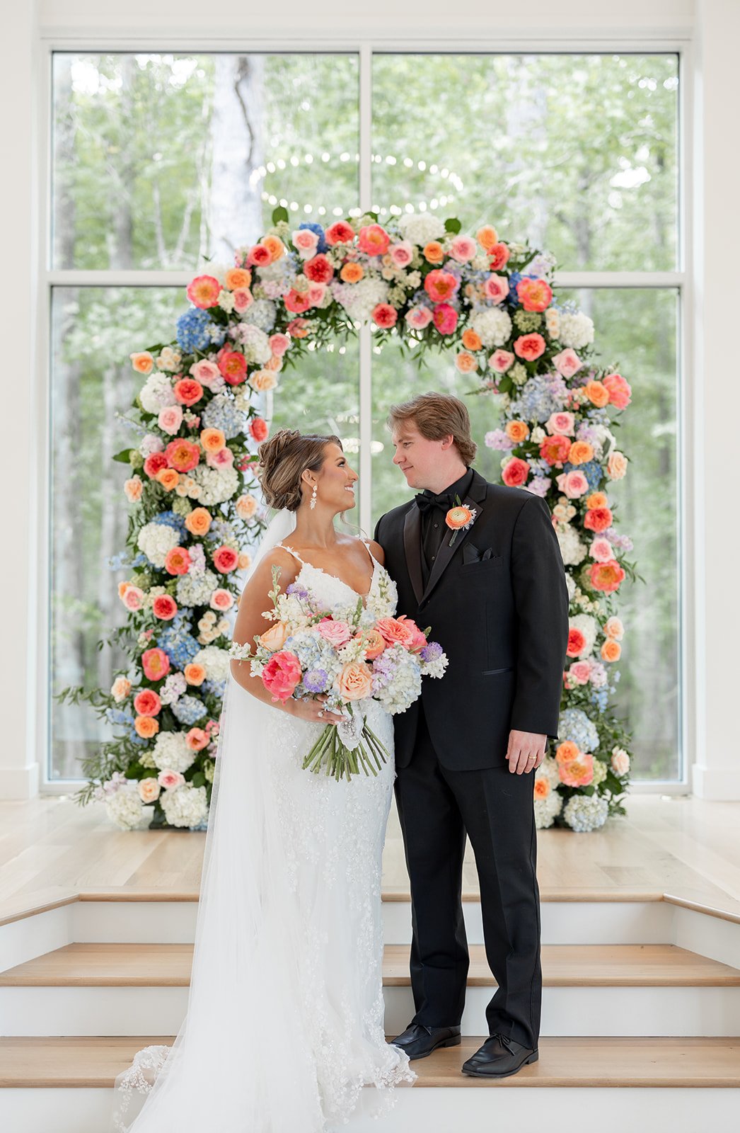 A bride and groom standing on a small stage facing each other, smiling, in front of a large window with a flower arch behind them, decorated with colorful flowers, during their wedding ceremony.