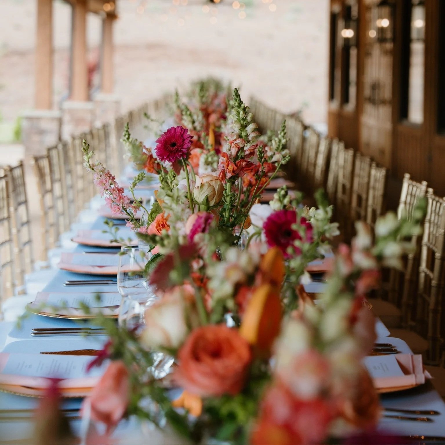 A long dining table decorated with a colorful floral centerpiece, set with plates, silverware, and wine glasses in a rustic indoor setting.