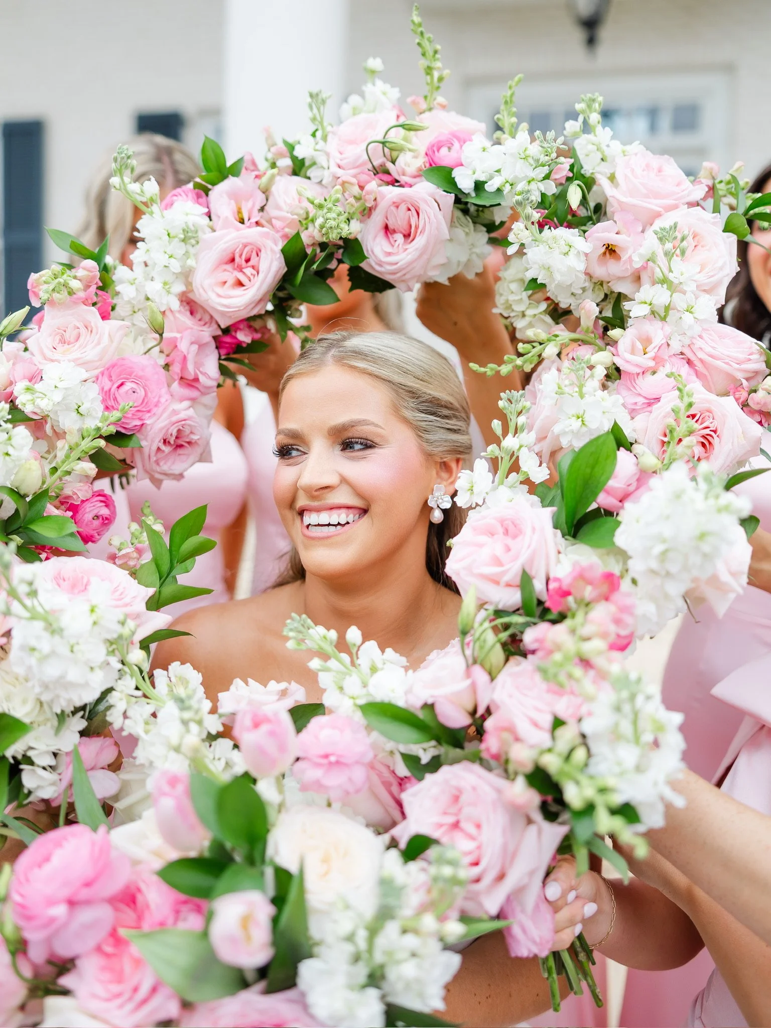 A woman with blonde hair and earrings is smiling amidst a large bouquet of pink and white flowers, possibly at a wedding or celebration.