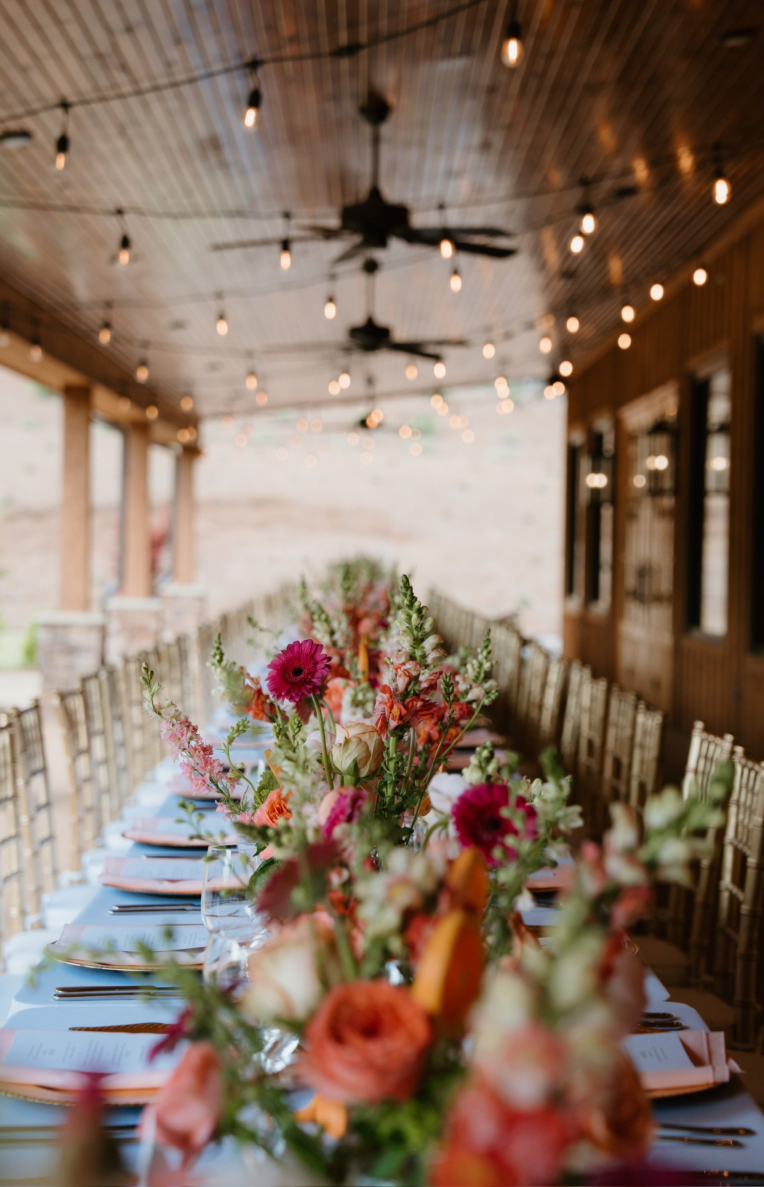 Blurred image of a long dining table decorated with a colorful floral centerpiece, set for an event, under a wooden ceiling with string lights and ceiling fans.