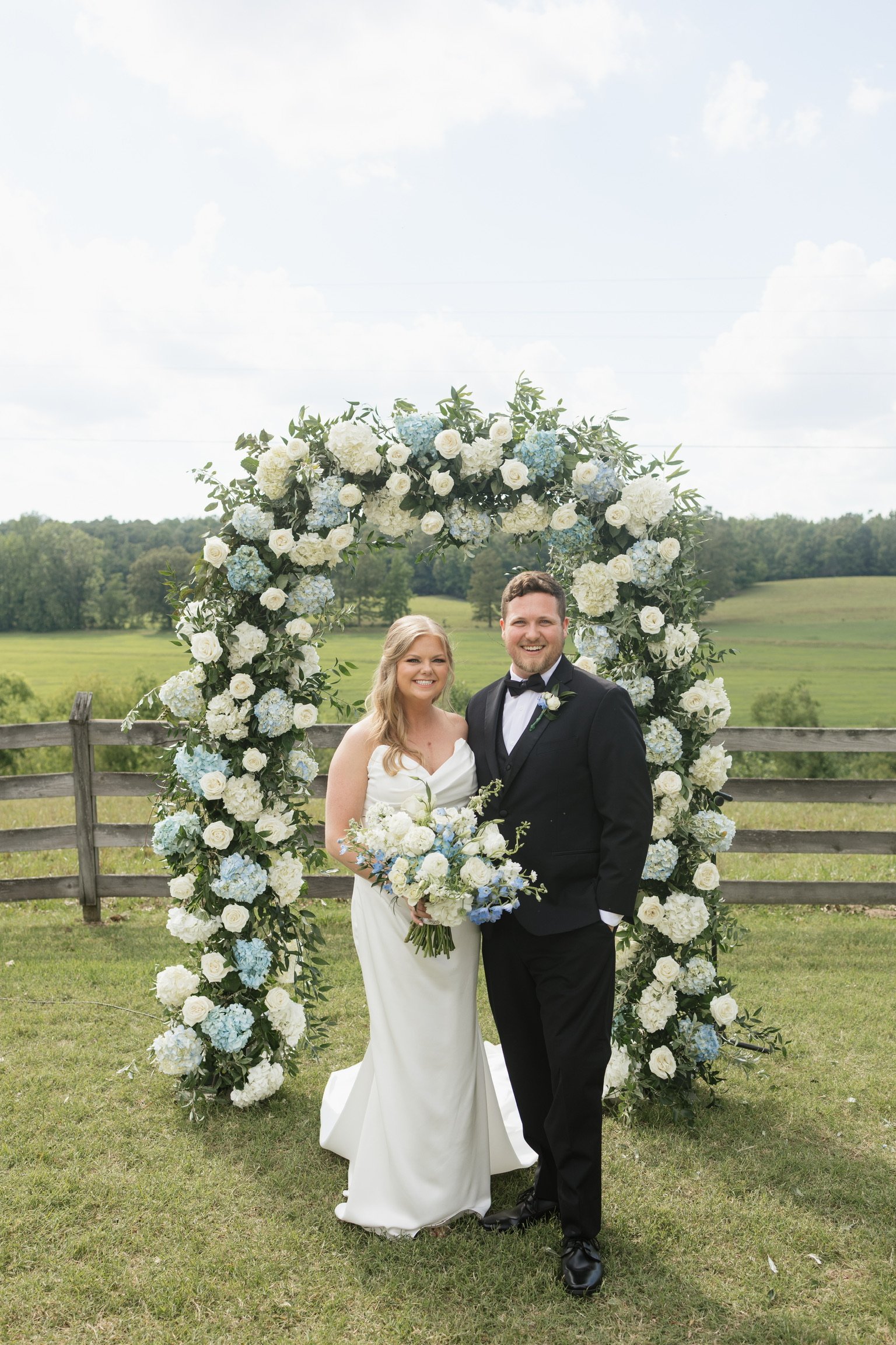 A bride and groom standing in front of a large circular floral arch at an outdoor wedding. The arch is decorated with white and light blue flowers and greenery. The background features a wooden fence and green open fields with trees under a partly cl