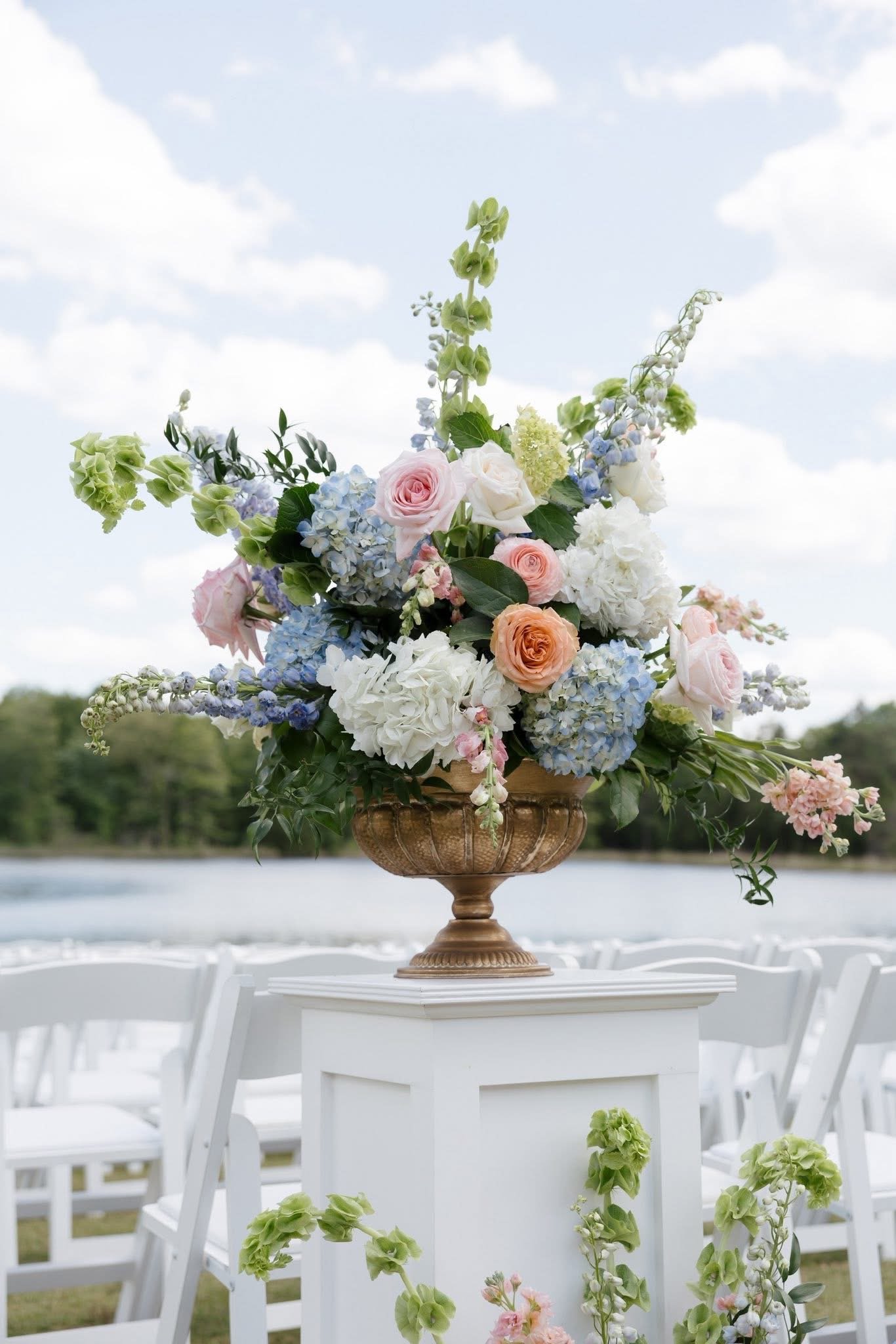 A floral arrangement featuring pink roses, white hydrangeas, blue delphiniums, and various greenery on a gold pedestal, outdoors near a body of water with a cloudy sky.