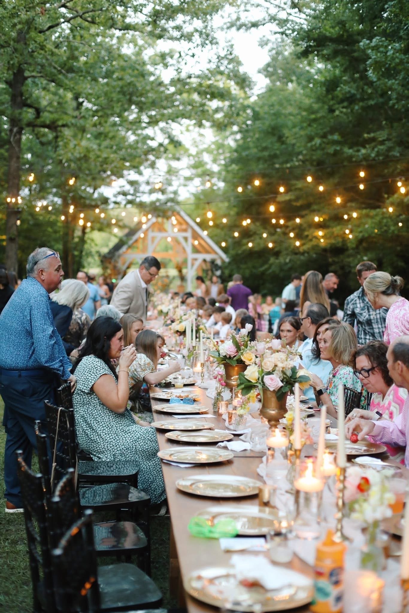 Outdoor wedding reception under string lights with long dining table decorated with flowers and candles, surrounded by guests.