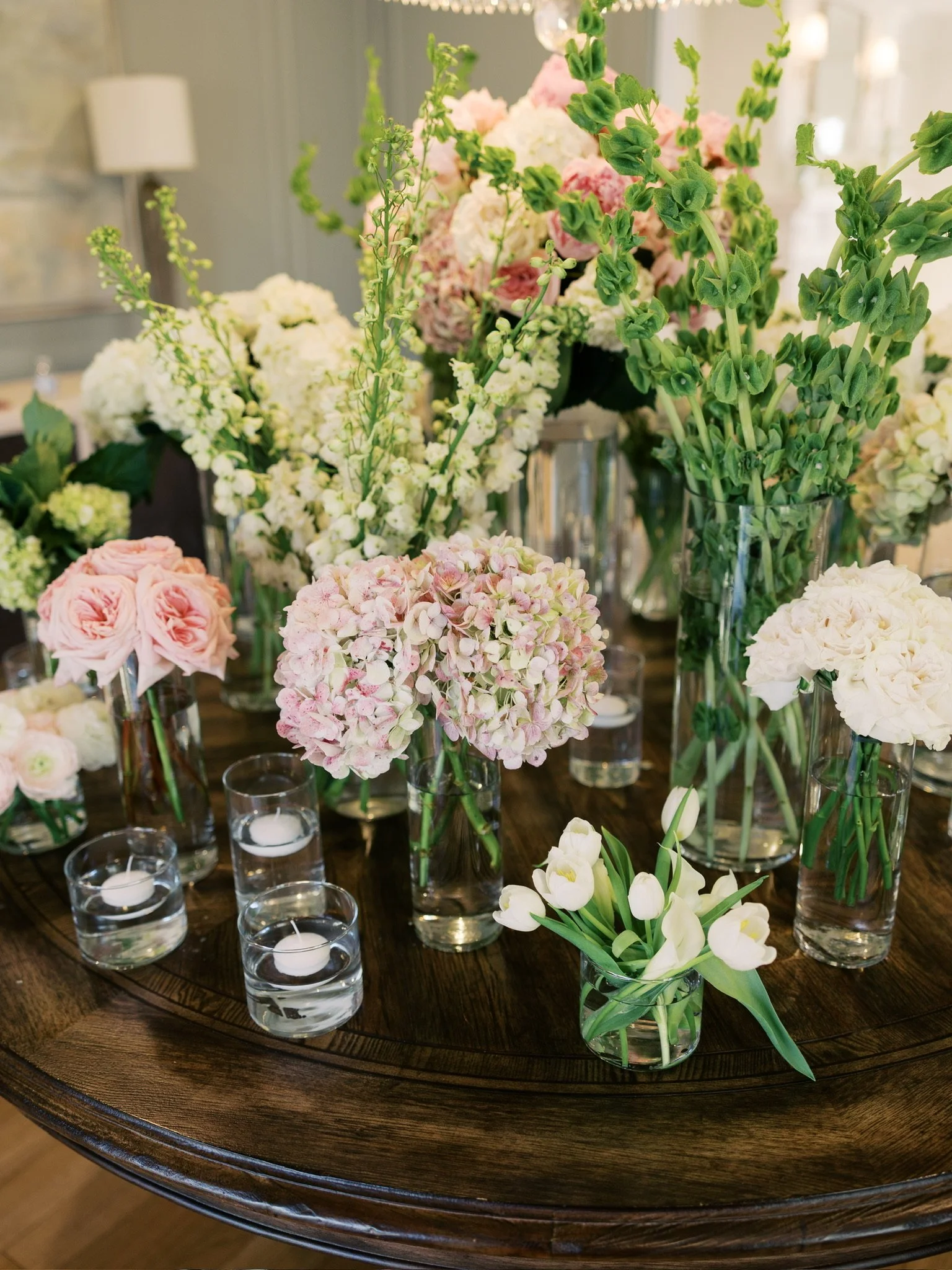 A dark wooden table with multiple clear glass vases filled with various flowers, including pink roses, white hydrangeas, and white calla lilies, arranged for decoration or event.
