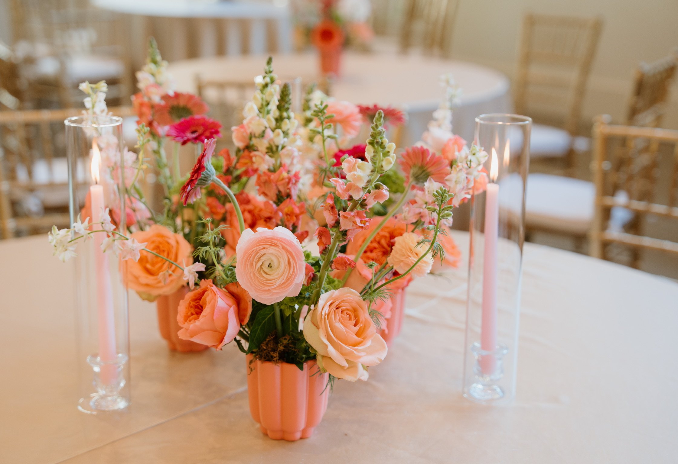 A floral centerpiece with pink, peach, and white flowers in a pink vase, surrounded by tall, clear glass candle holders with lit candles on a table.