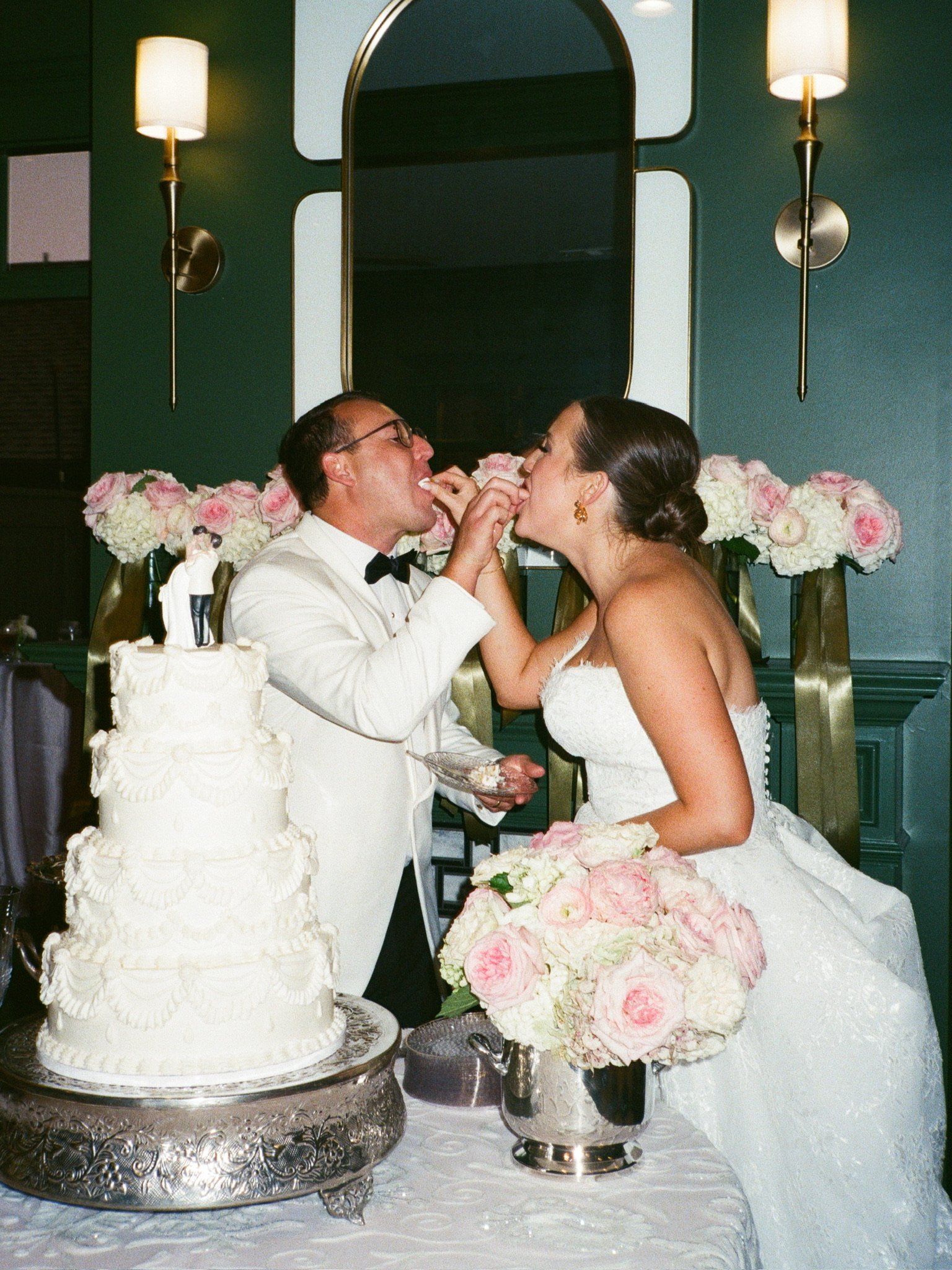 A bride and groom are feeding each other cake at their wedding reception. There is a decorated wedding cake with several tiers and intricate frosting details. Floral arrangements with pink and white flowers are behind them, and a mirror, wall sconces