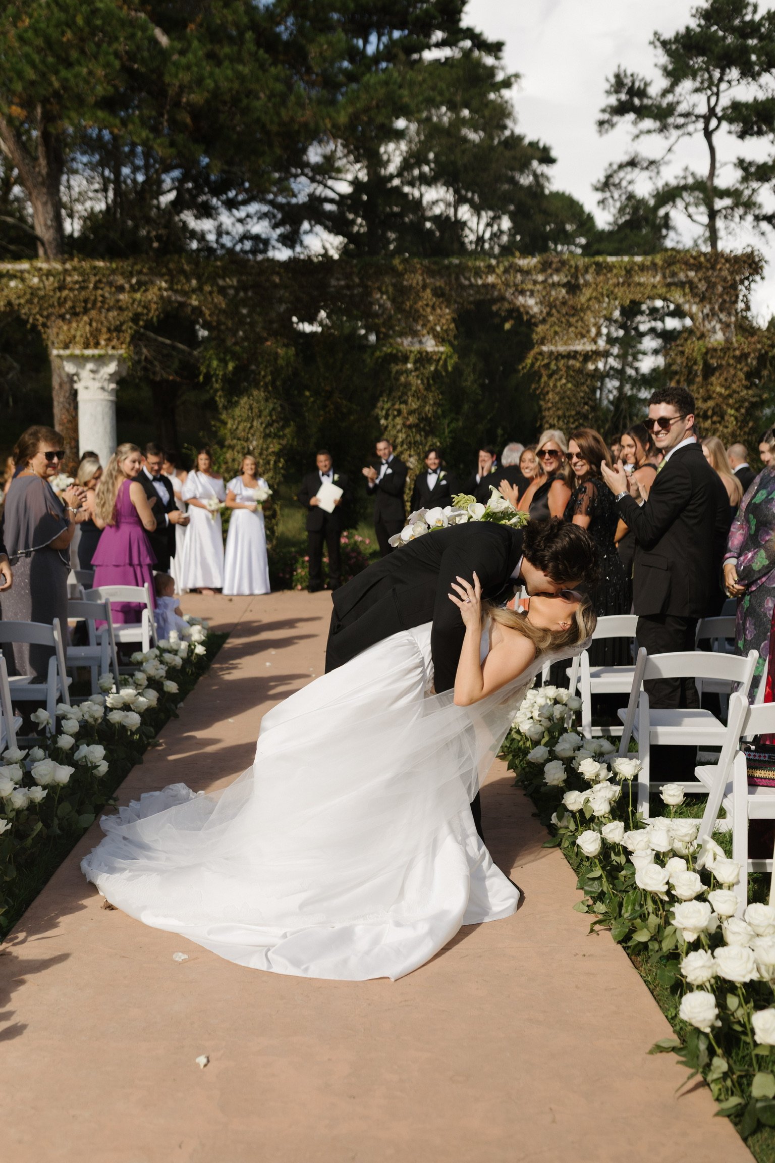 A bride and groom share a kiss during their outdoor wedding ceremony, with guests standing and clapping in the background, amid white floral decorations and a green, scenic backdrop.