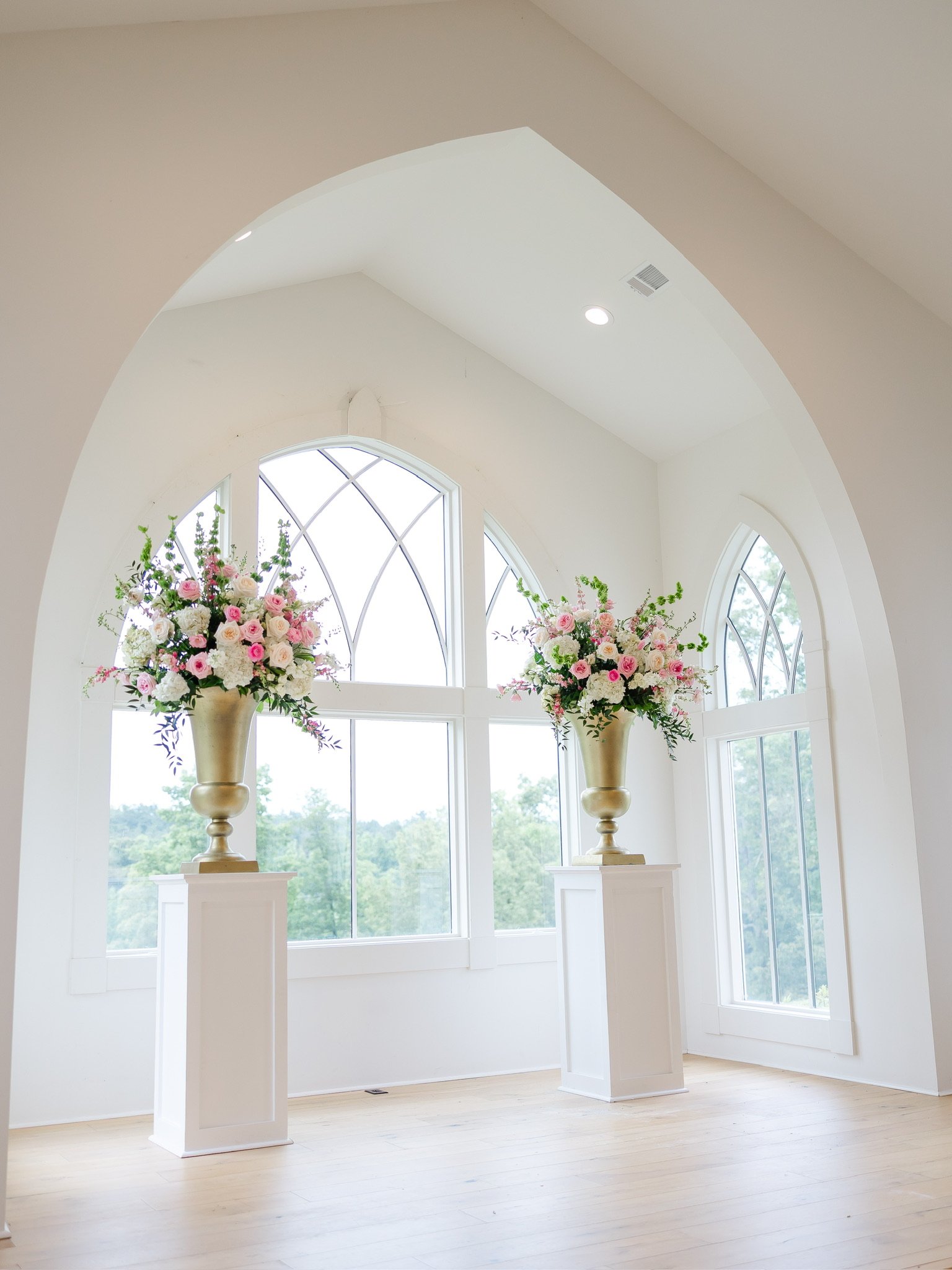 A room with large arched windows and two tall floral arrangements in gold vases on white pedestals.