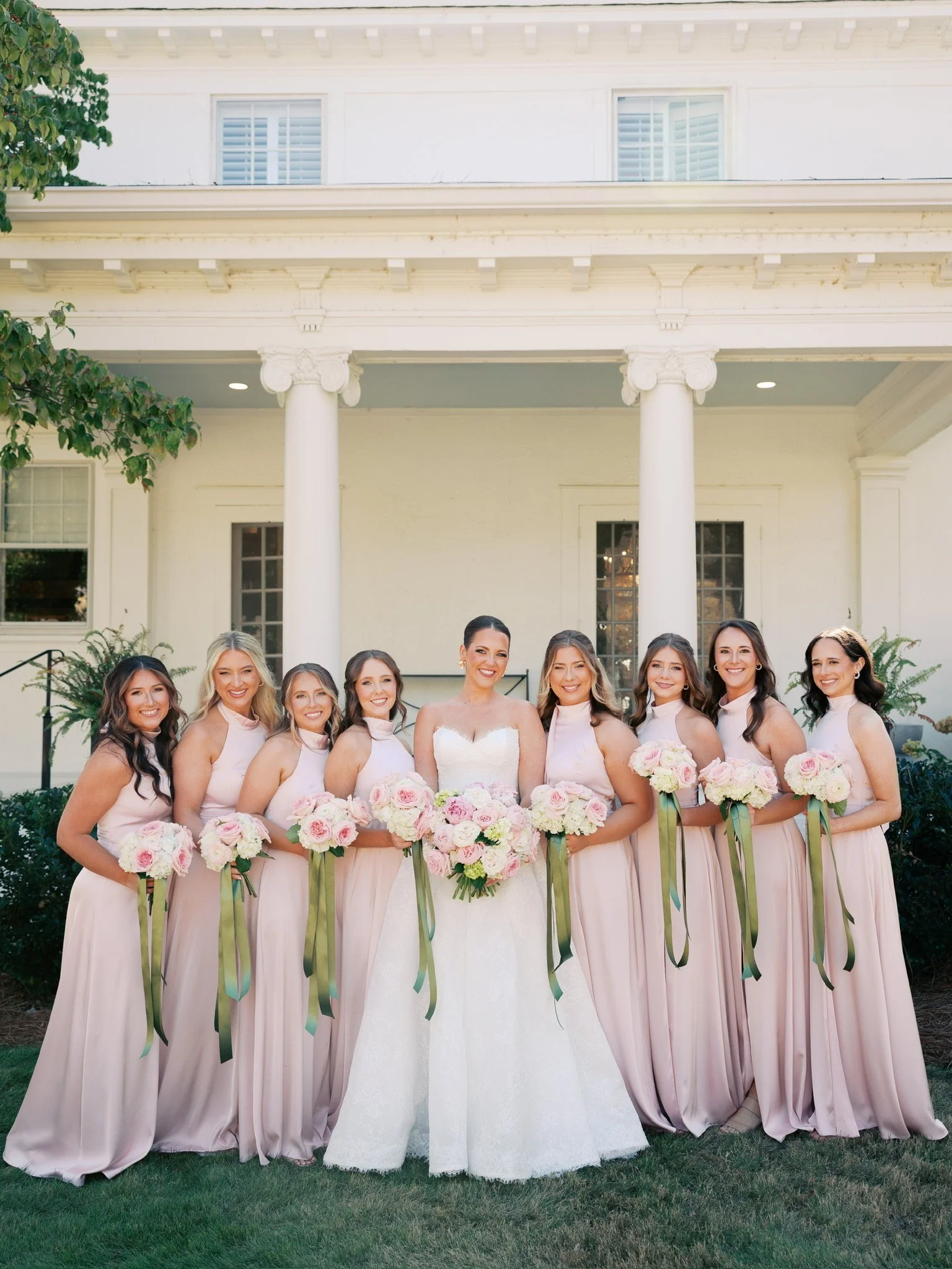 A bride and her bridesmaids standing in front of a white house with columns, holding bouquets of pink and white roses with green ribbons.