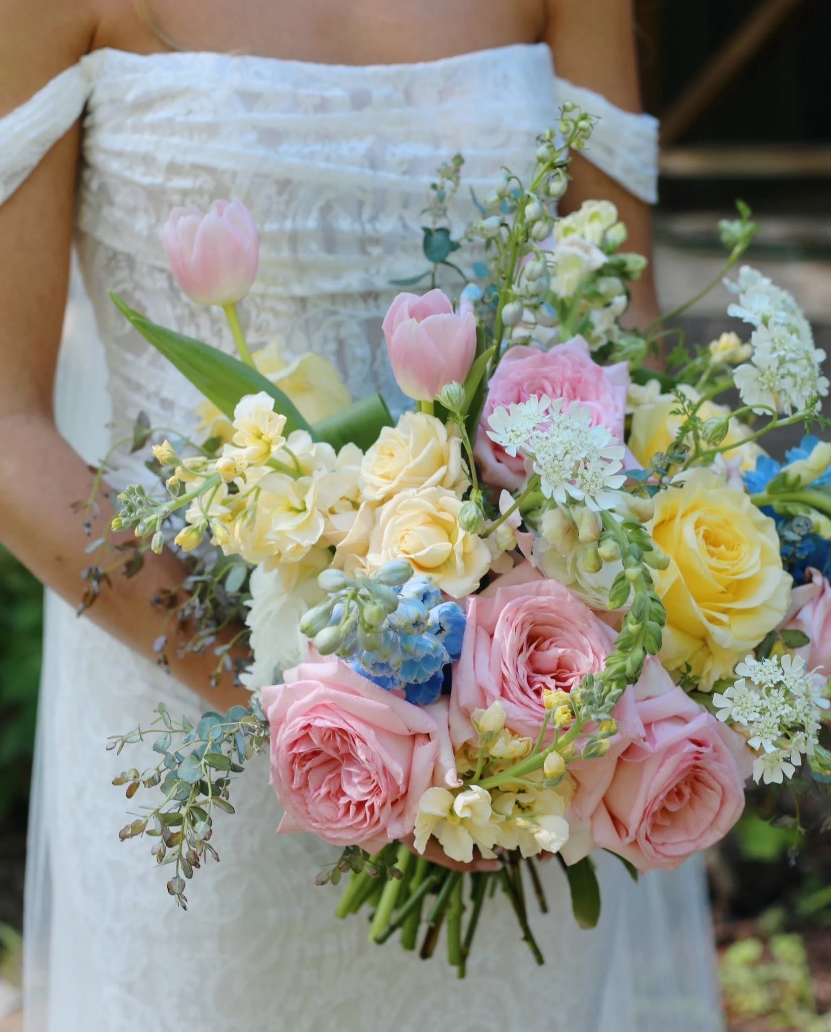 A person holding a bouquet of pink, yellow, and white flowers, including tulips, roses, and other blossoms, with a white dress in the background.