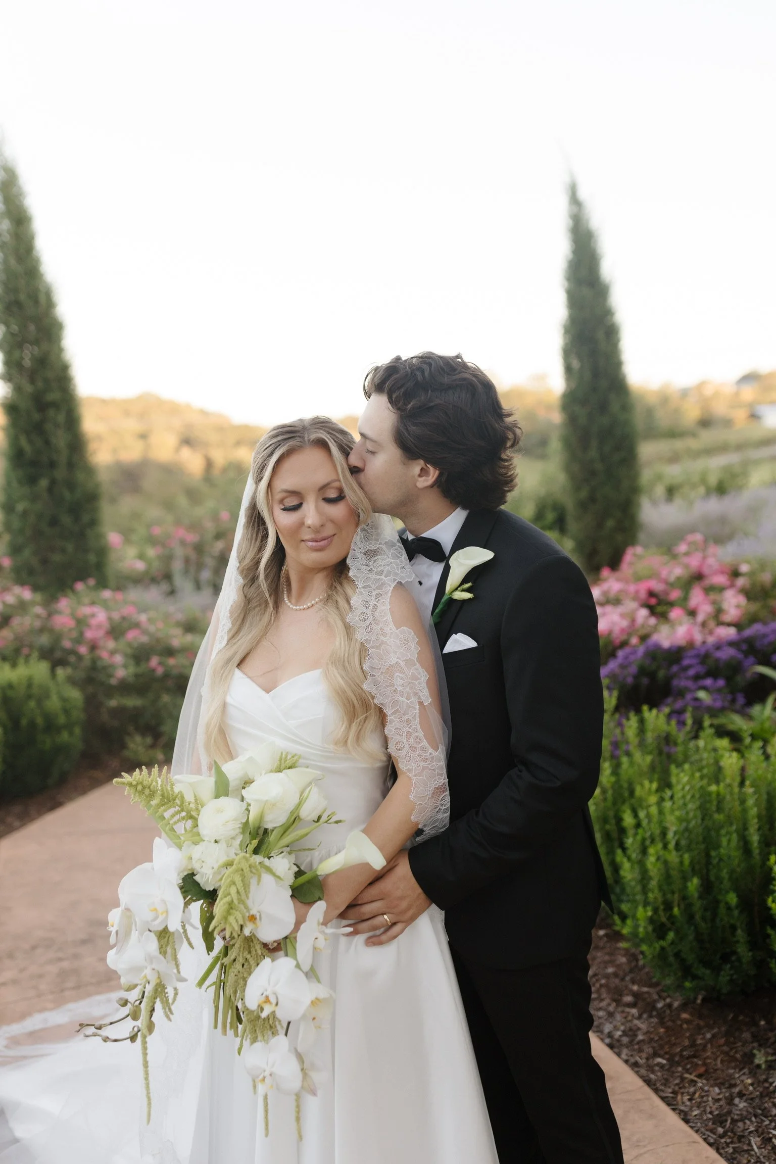 A bride and groom standing outdoors during their wedding ceremony. The groom is kissing the bride on the temple. The bride is holding a bouquet of white flowers and is wearing a wedding dress with lace details and a ribbon. The groom is dressed in a 