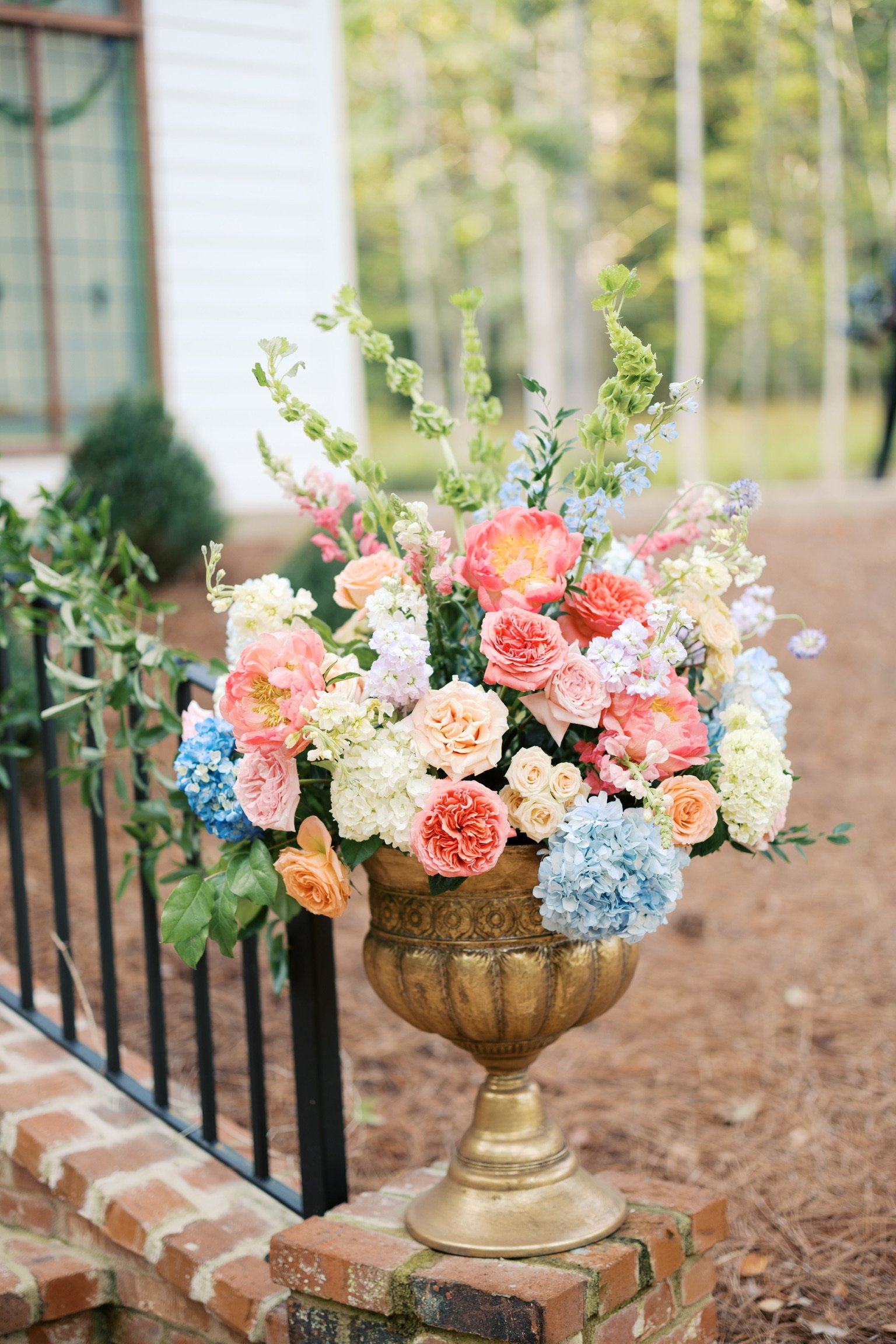 A large floral arrangement in a gold ornate vase, featuring pink, peach, blue, white, and light purple flowers, placed outdoors on a brick and mortar surface.