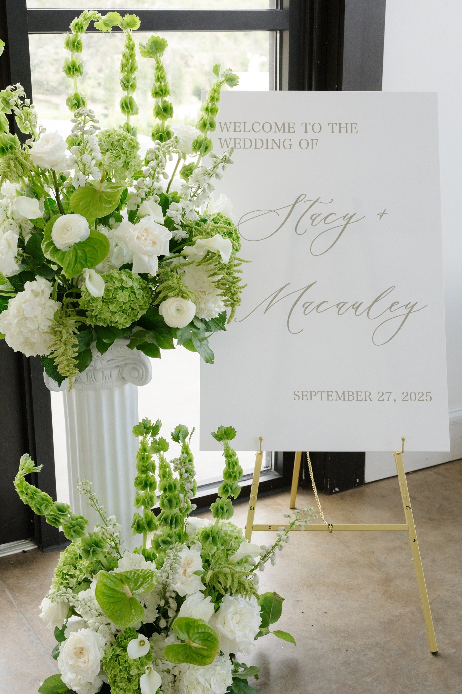 White floral arrangement next to a wedding welcome sign with green and white flowers and text for a wedding on September 27, 2025.