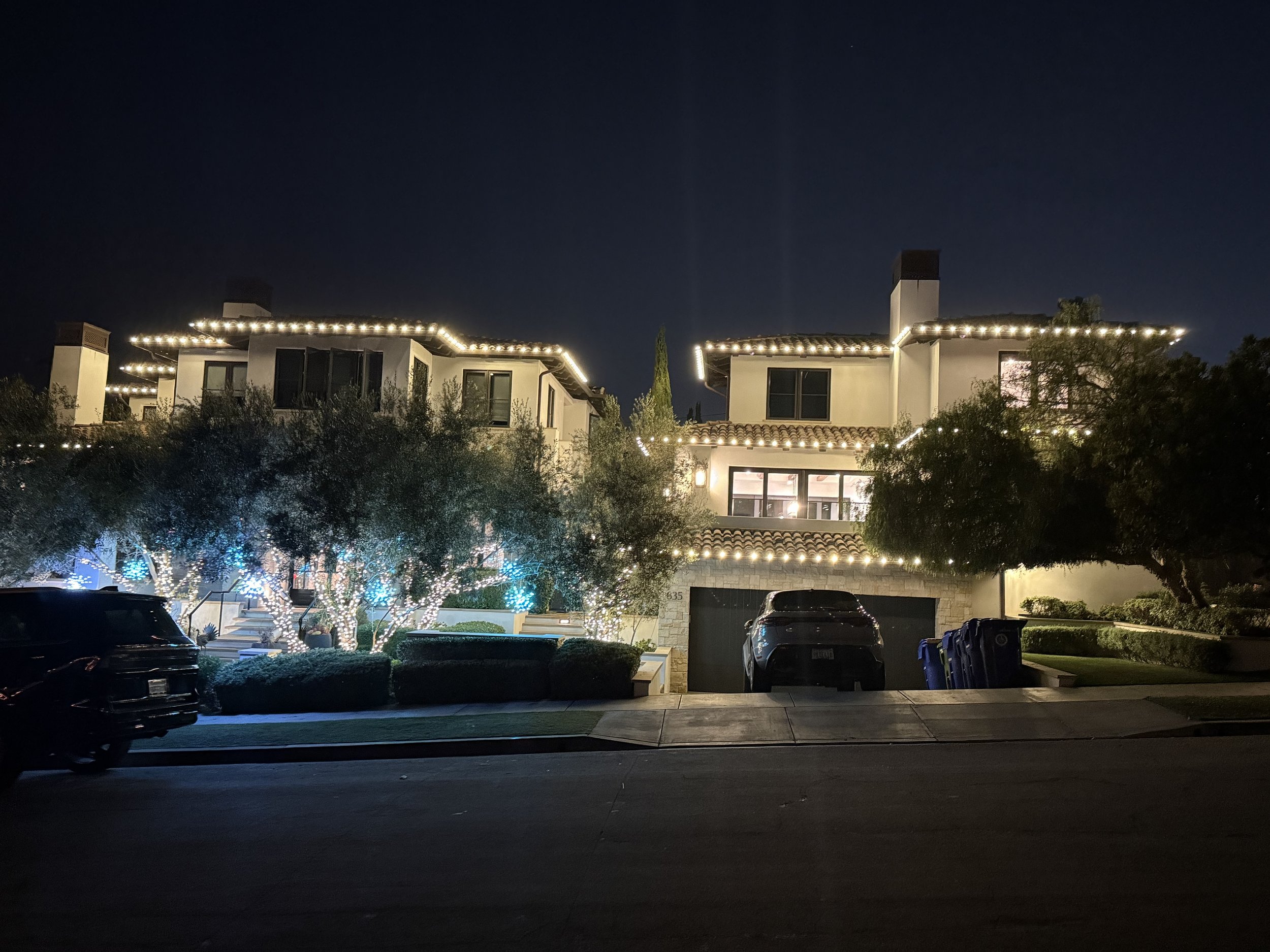 Nighttime view of a residential house decorated with string lights outlining the roof and upper balcony. The house has multiple chimneys, large windows, and a surrounding garden with lit trees and bushes. There are parked cars in the driveway.