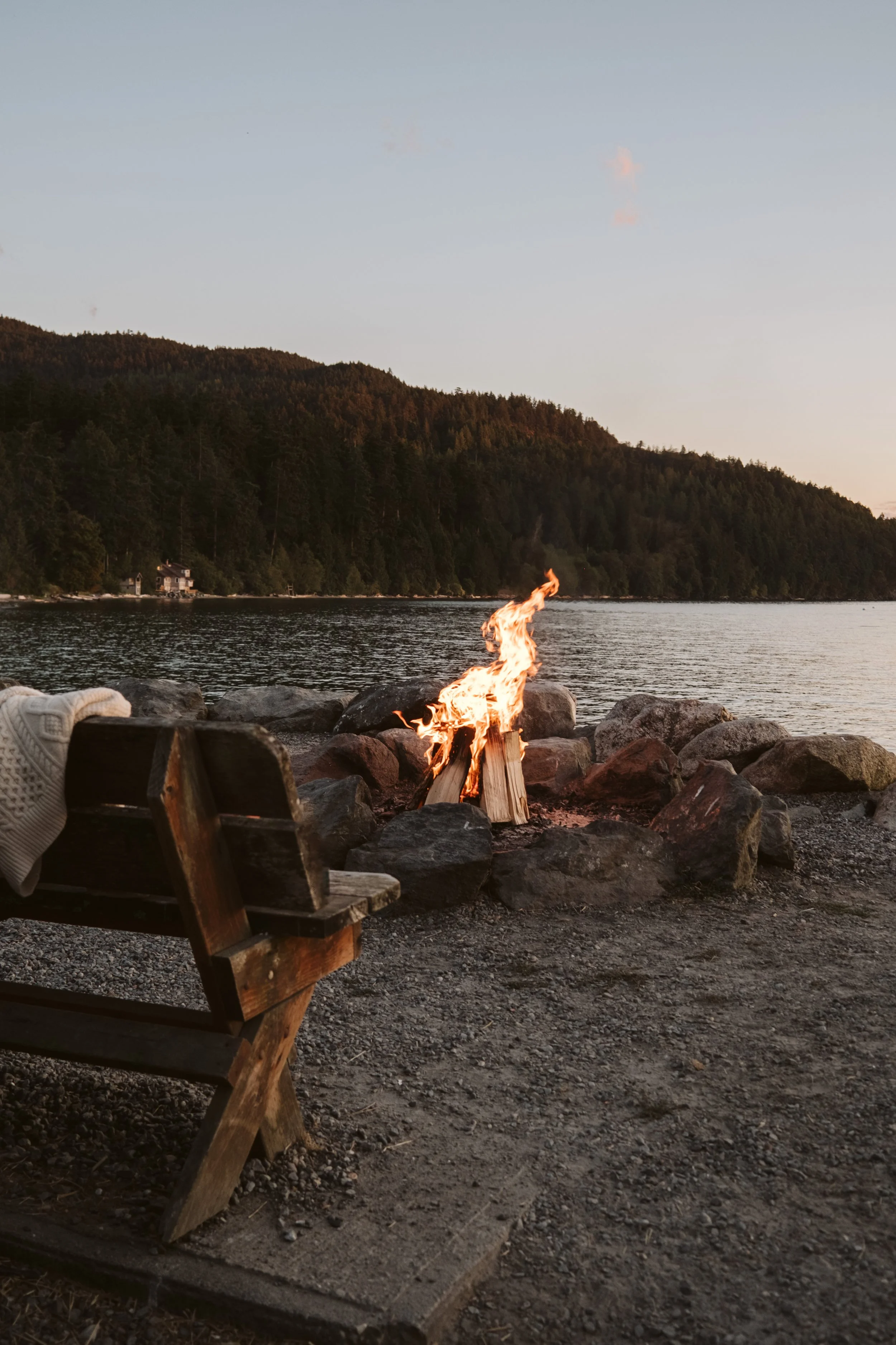 A campfire on a rocky beach near a lake, with a hill covered in trees and a house visible in the background during sunset.