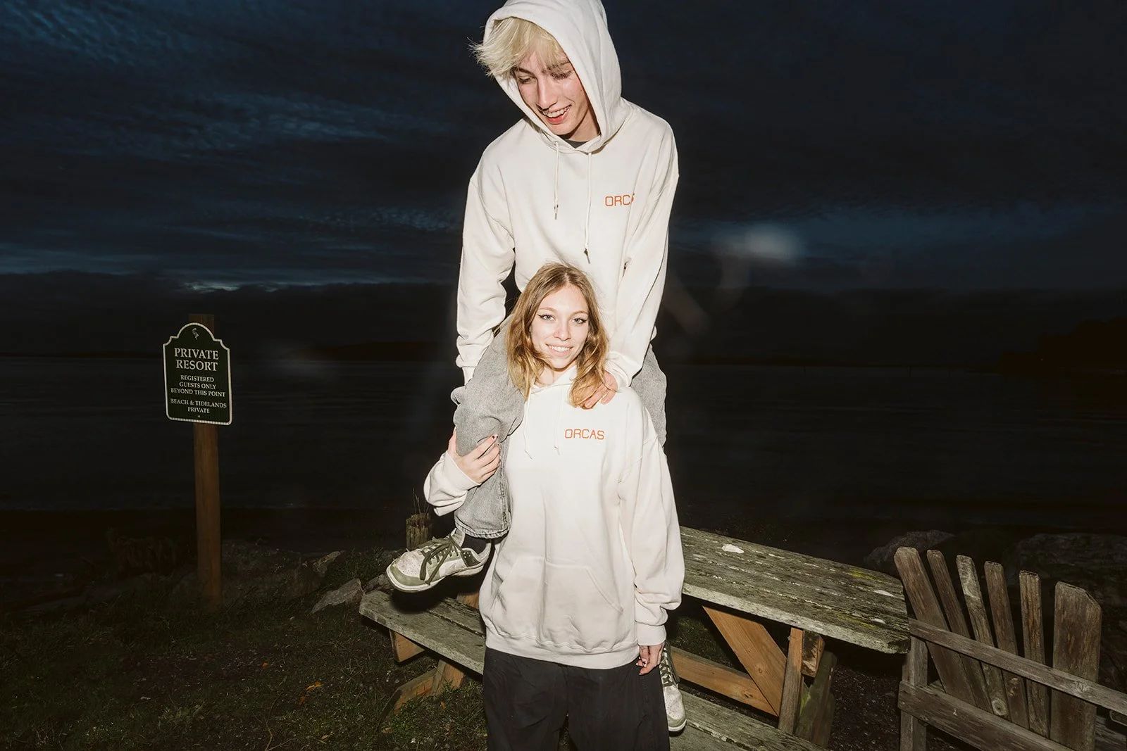 Two young women in white hoodies with 'ORCAS' logos on the front, one standing and the other sitting on the other's shoulders, smiling at the camera, outdoors at a dark beach or shoreline at dusk, with a sign that reads 'Private Resort' in the background.