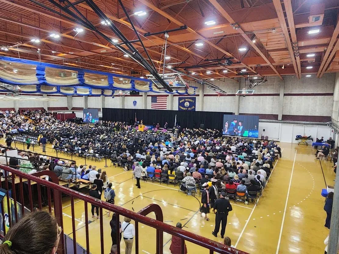 Graduation ceremony in a gymnasium, with many people seated and graduates in caps and gowns. There are large screens on stage showing speakers, an American flag, and banners for a community college.