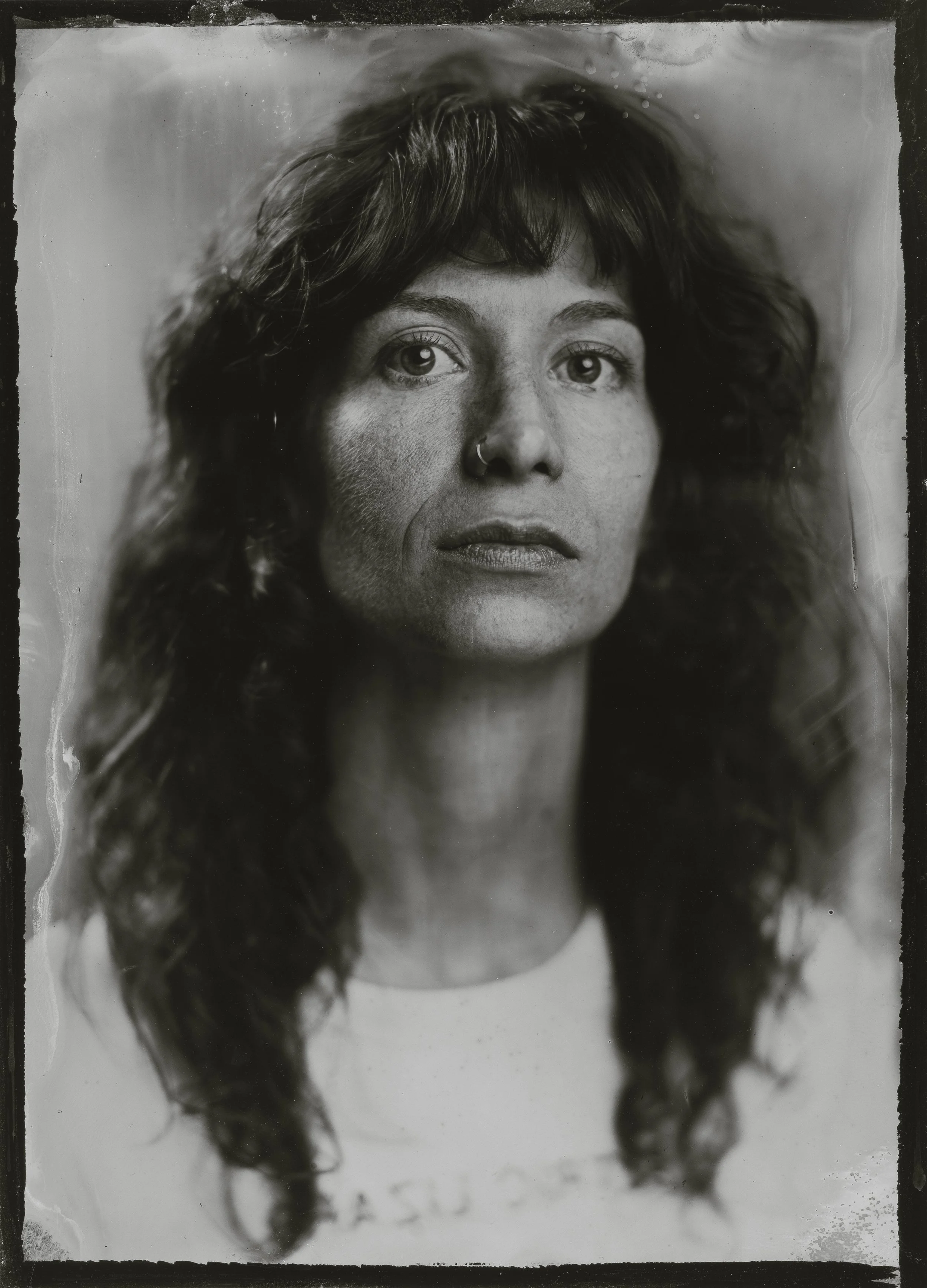 Black and white portrait of a woman with wavy hair, wearing a nose ring and a white shirt.