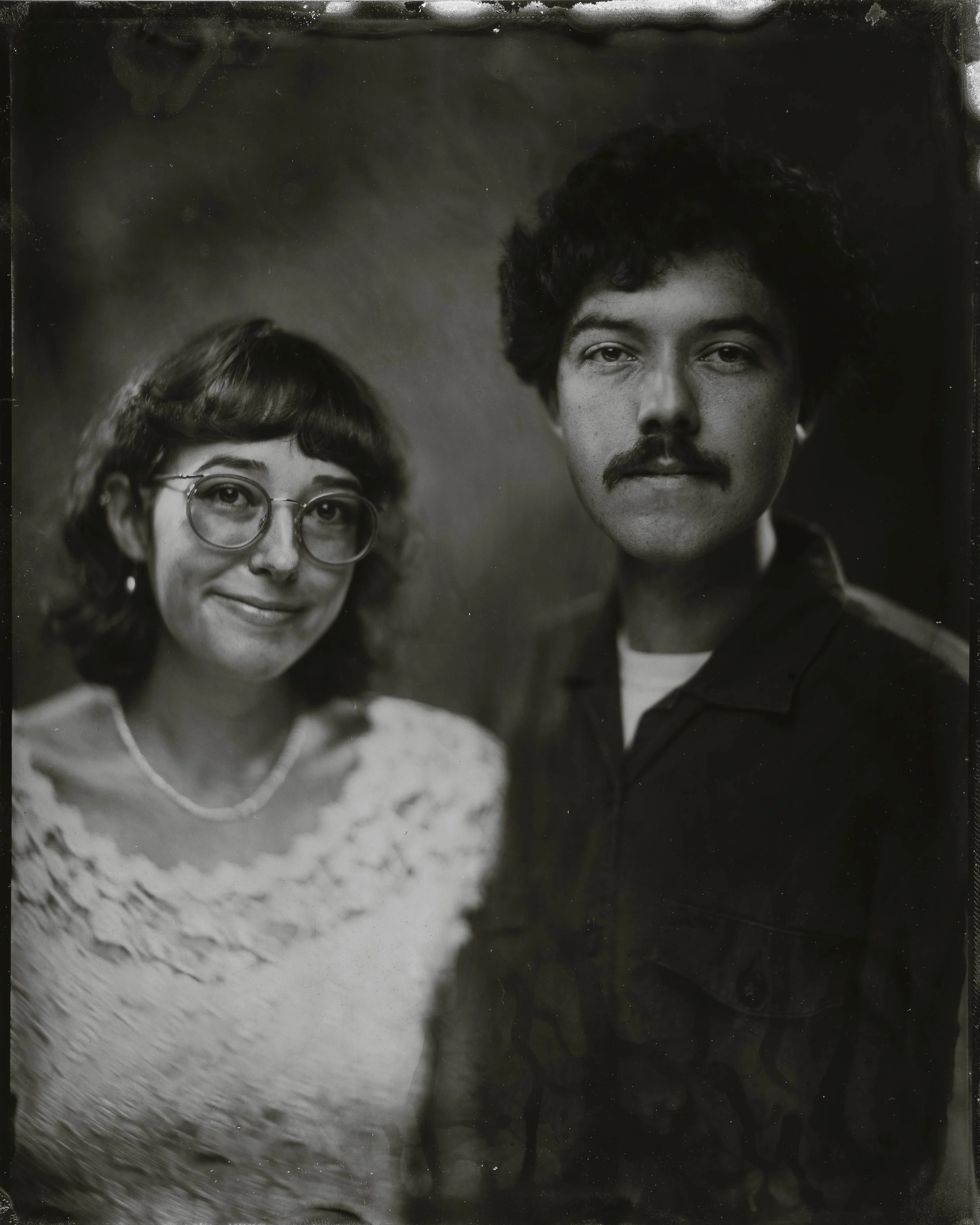 A black-and-white portrait of a woman with glasses and a man with a mustache, both looking at the camera.