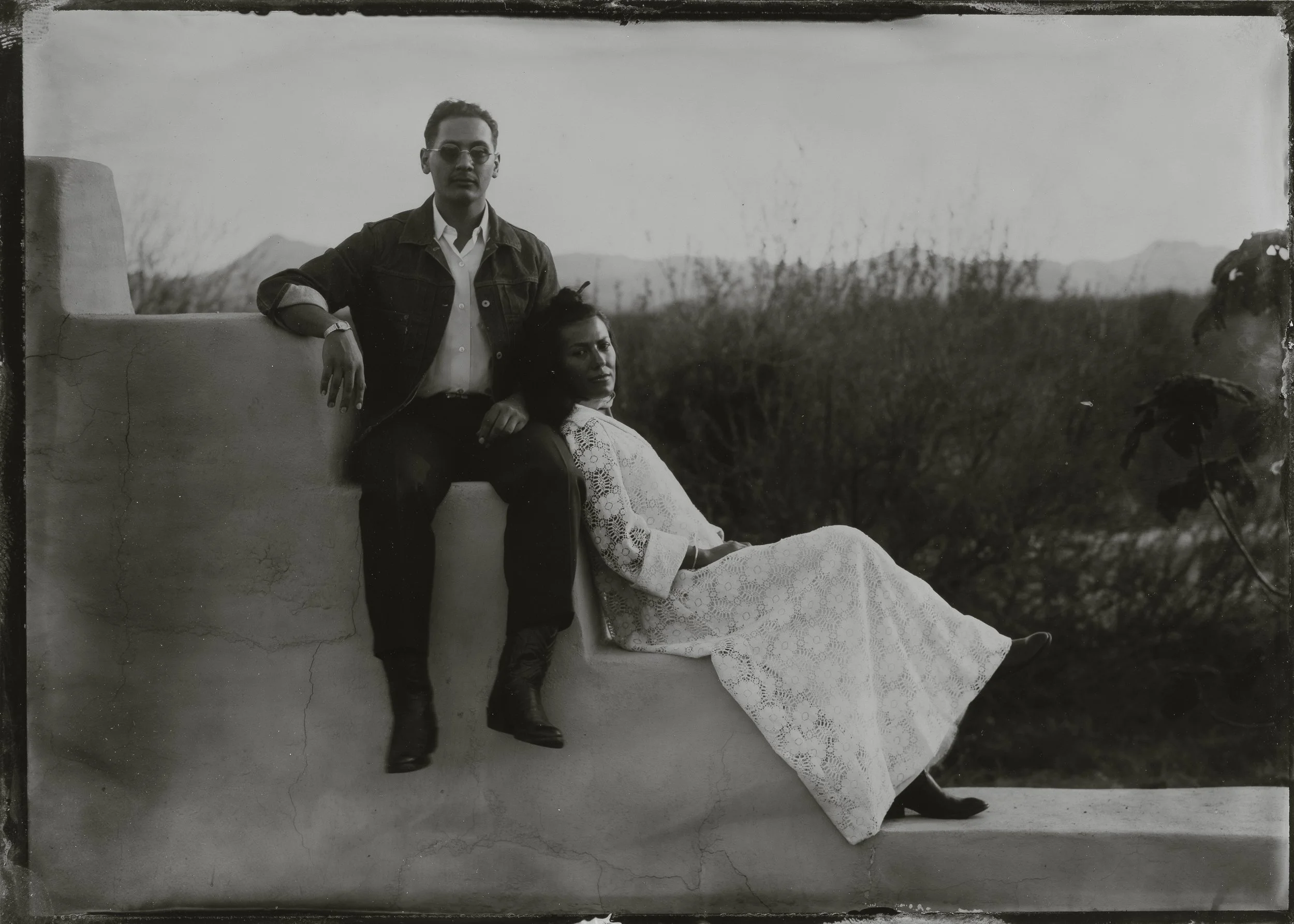 A black and white photo of a man and woman outdoors. The man is standing with his arm resting on a wall, wearing glasses, a denim jacket, and is dressed in a shirt and pants. The woman is sitting on the wall, leaning back with her legs extended, wear