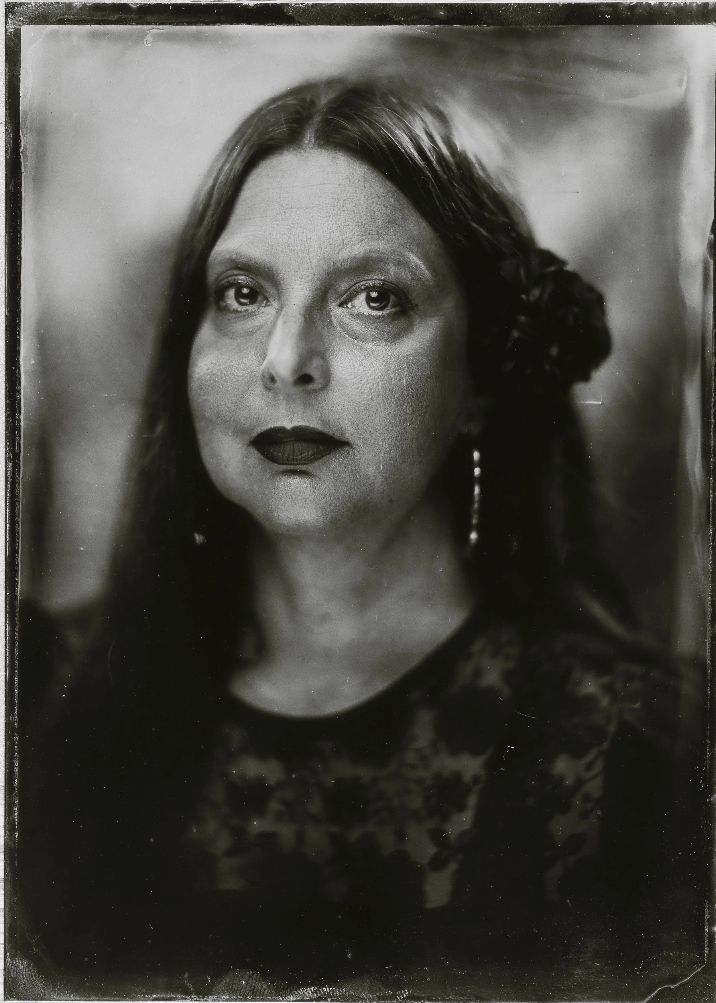 Black and white portrait of a woman with long dark hair, dark lipstick, and earrings, looking directly at the camera.
