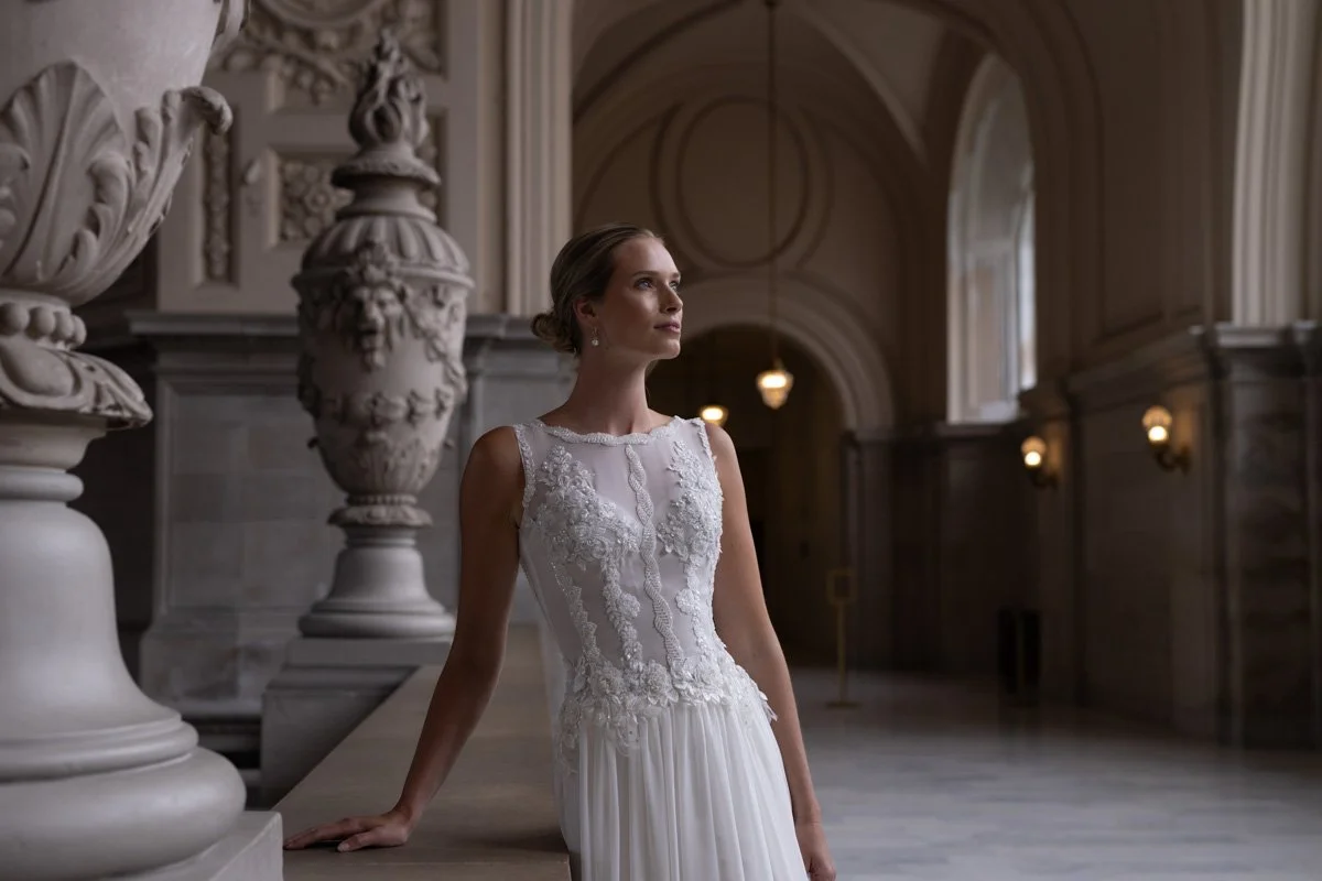 Photograph of a model wearing a sleeveless wedding gown, posing in a large marble hall.