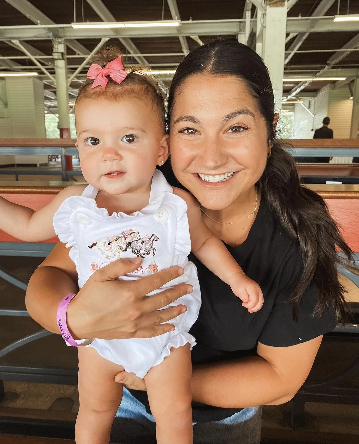 Smiling woman holding a baby wearing a white outfit with a pink bow, indoors.