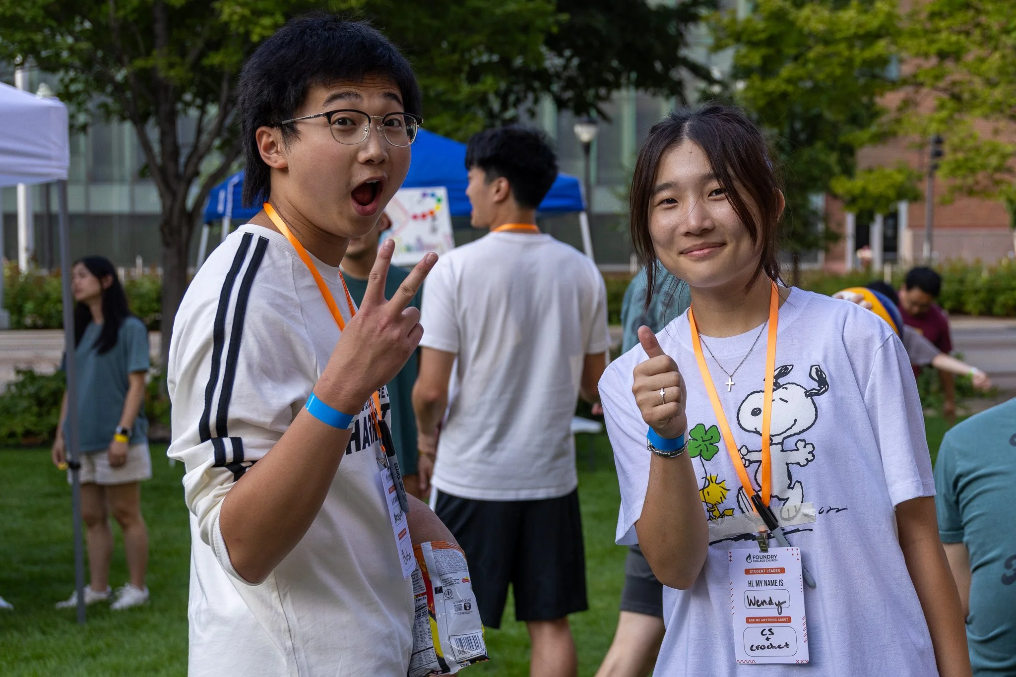 Two teenagers smiling and posing for a photo outdoors at an event, with attendees and trees in the background.