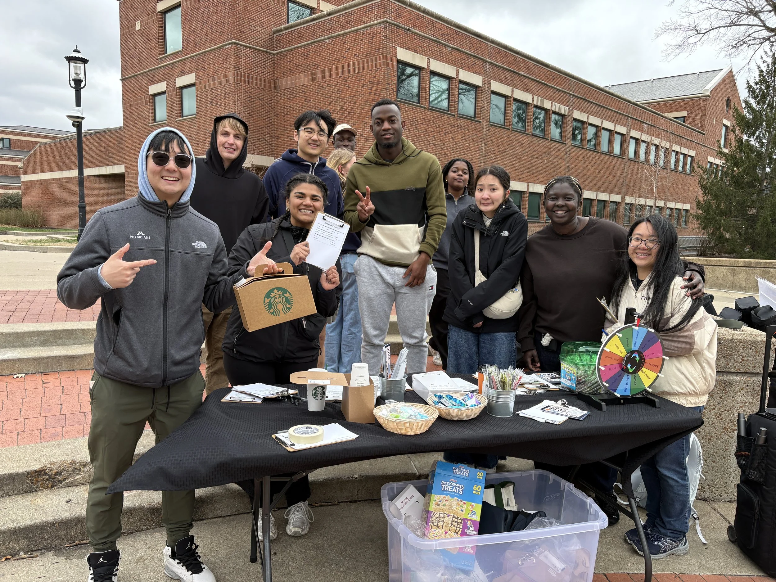 A diverse group of people gathered behind a table outdoors in front of a brick building. The table has various items including a colorful wheel, treats in a box, and flyers. The group is smiling and posing for the photo, with some making gestures like a peace sign.