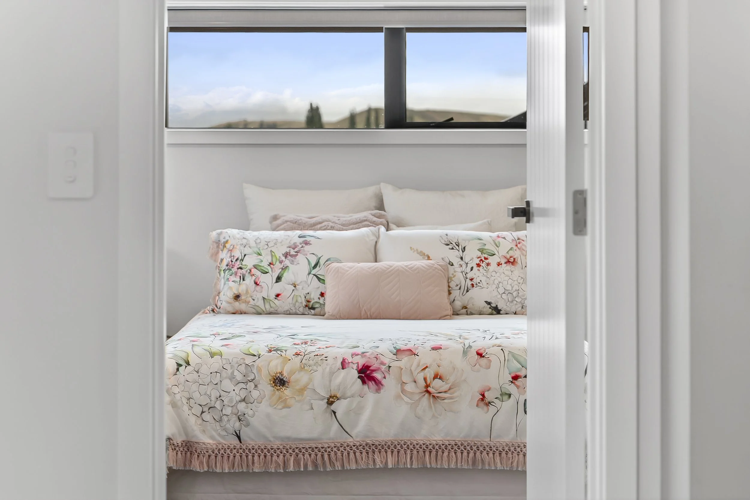 A bedroom with a window showing a view of hills and sky. The bed has floral bedding, including a white duvet with colorful flower prints, and multiple pillows in neutral tones.