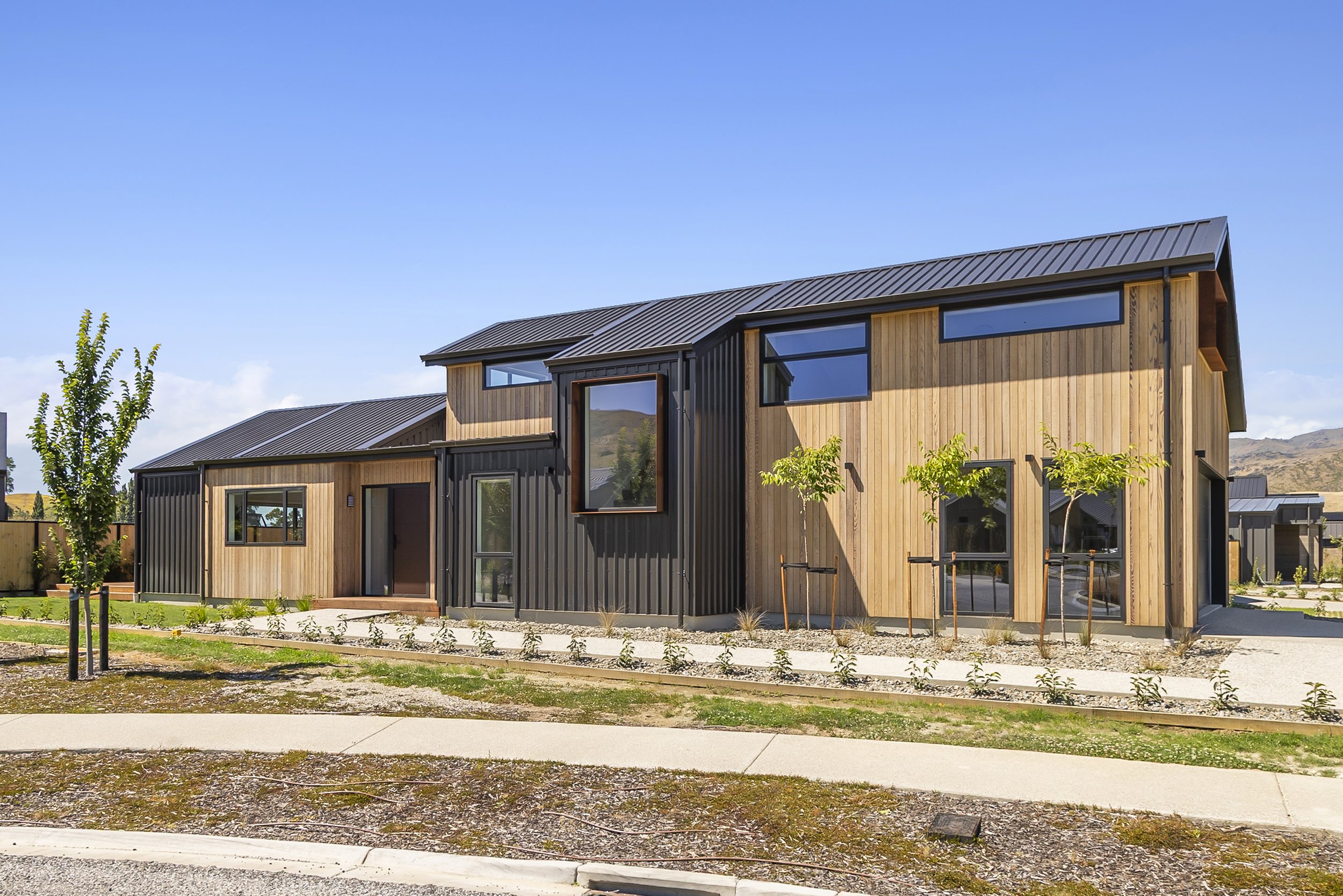 Modern residential building with wooden and black metal exterior, multiple large windows, small trees, and a clear blue sky in the background.