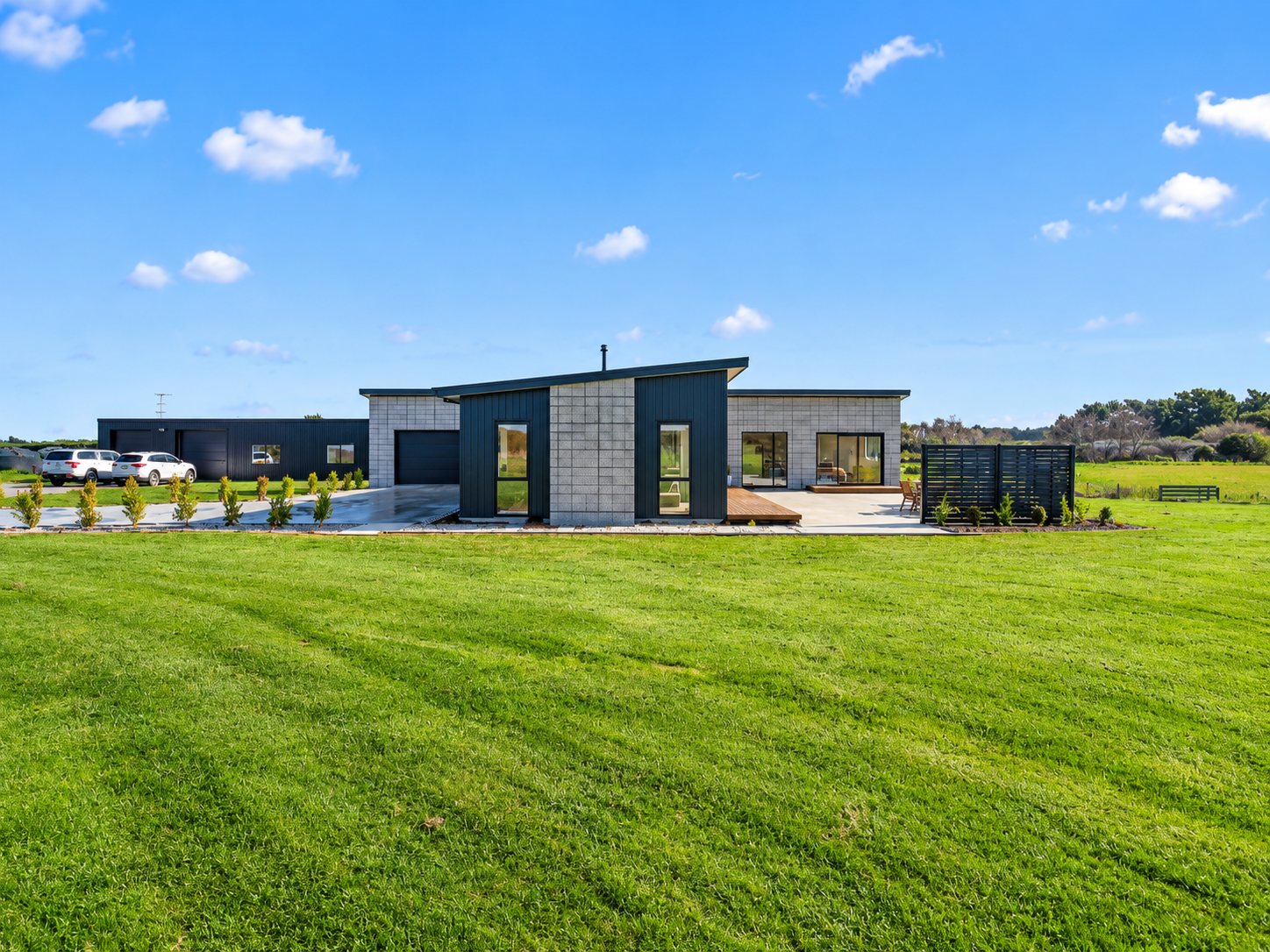 Modern house with gray concrete and dark blue siding, large windows, and a spacious green lawn, under a bright blue sky with scattered clouds.